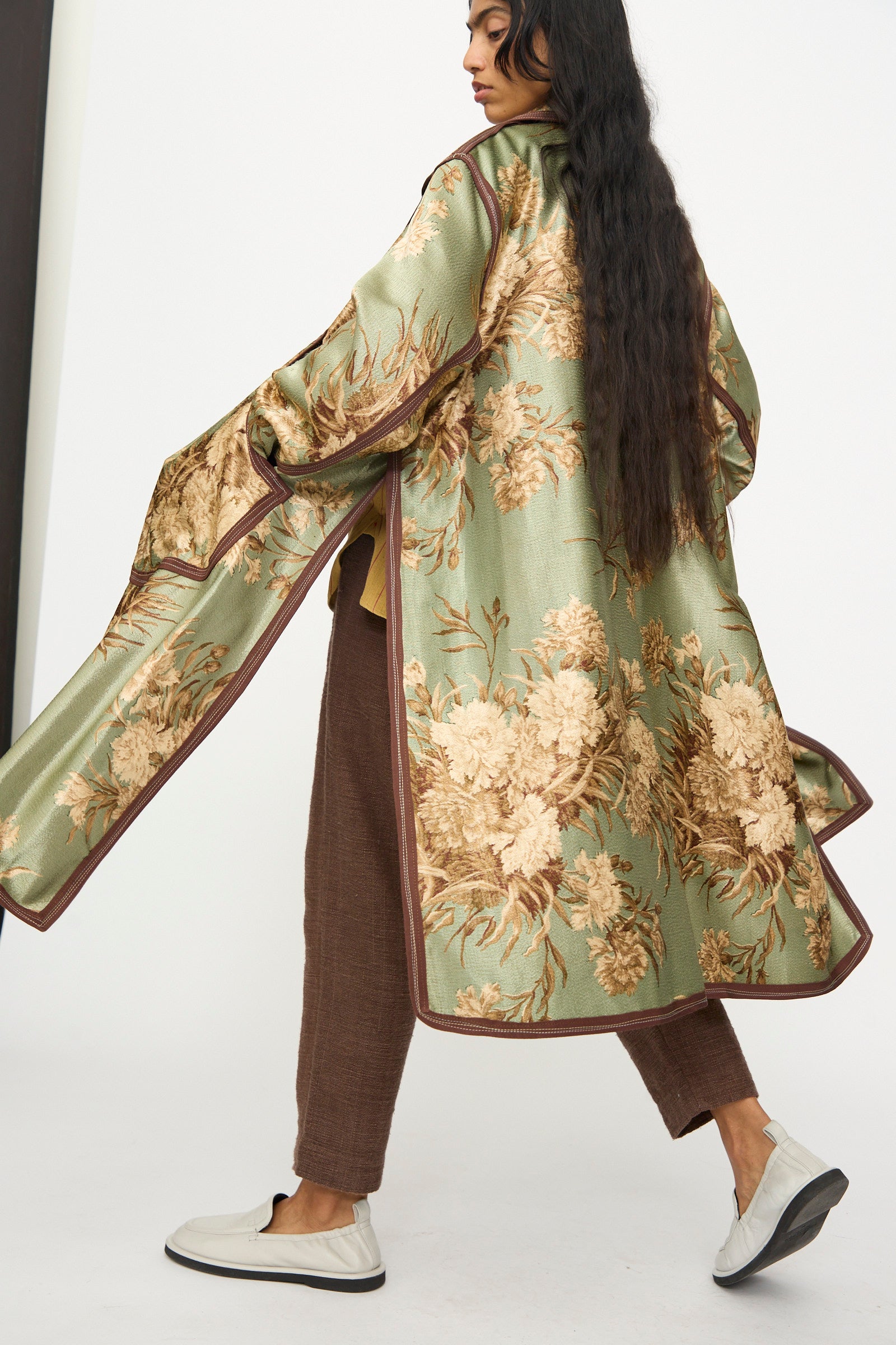 A person in the Thank You Have A Good Day Taped Trench in Floral, brown pants, and white slip-on shoes stands against a plain background, facing slightly away from the camera.