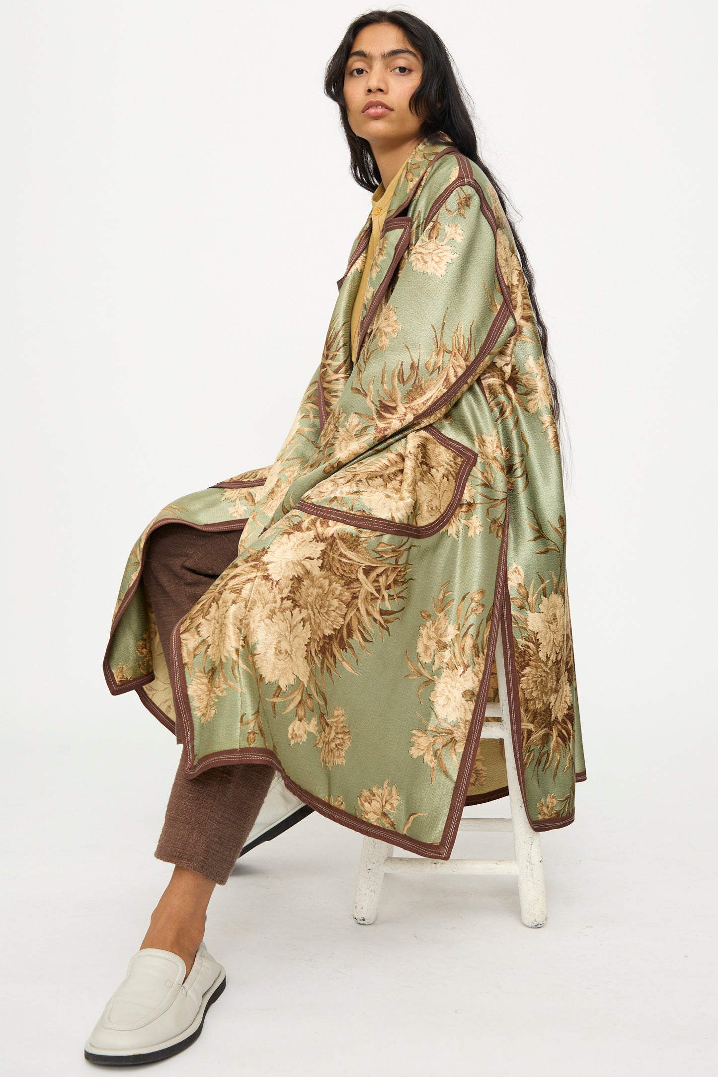 A person sits on a stool against a white background, wearing the Thank You Have A Good Day Taped Trench in Floral, beige-toned with green flowers, paired with brown pants and white slip-on shoes.