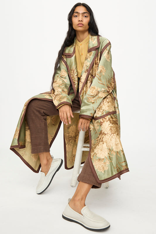 A person with long dark hair sits on a white stool, wearing the Thank You Have A Good Day Taped Trench in Floral, brown pants, a yellow top, and white slip-on shoes against a plain white background.