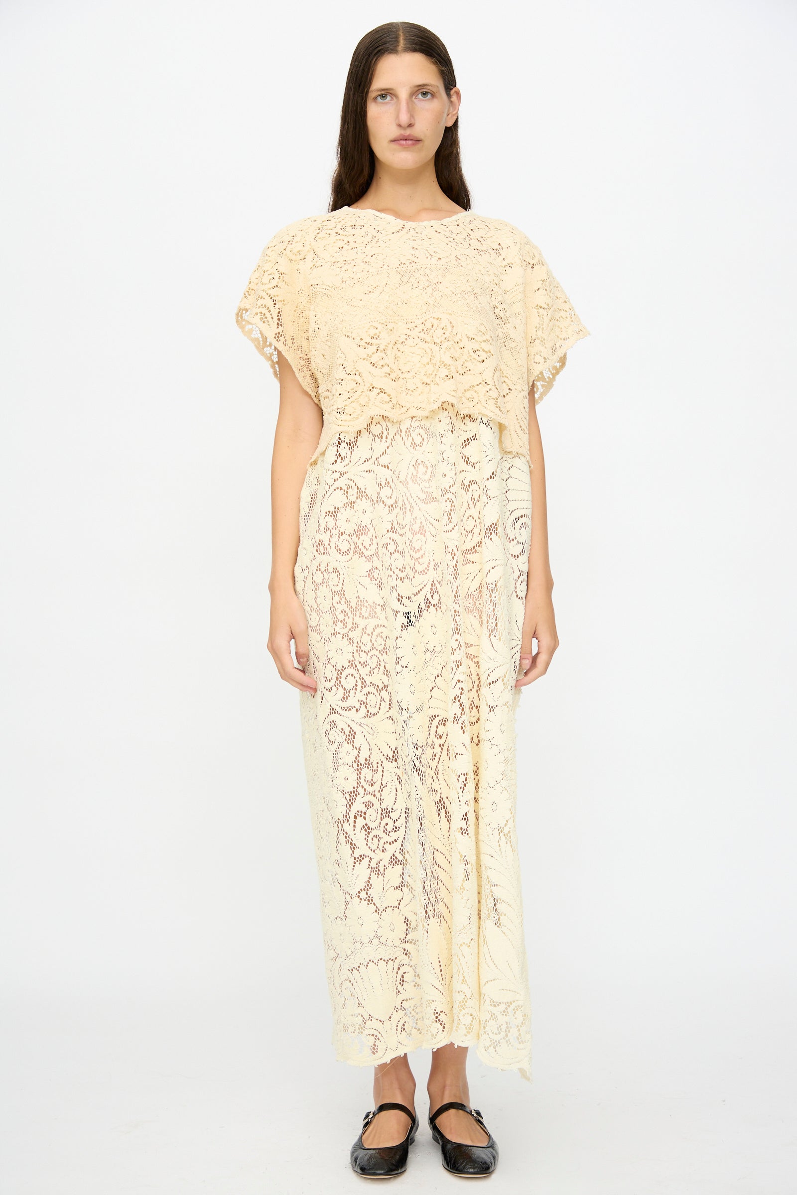 A woman faces forward in the Tilda Gown in Quaker Lace by Thank You Have A Good Day, a sheer cream maxi dress with short sleeves, paired with black flats, set against a plain white background.