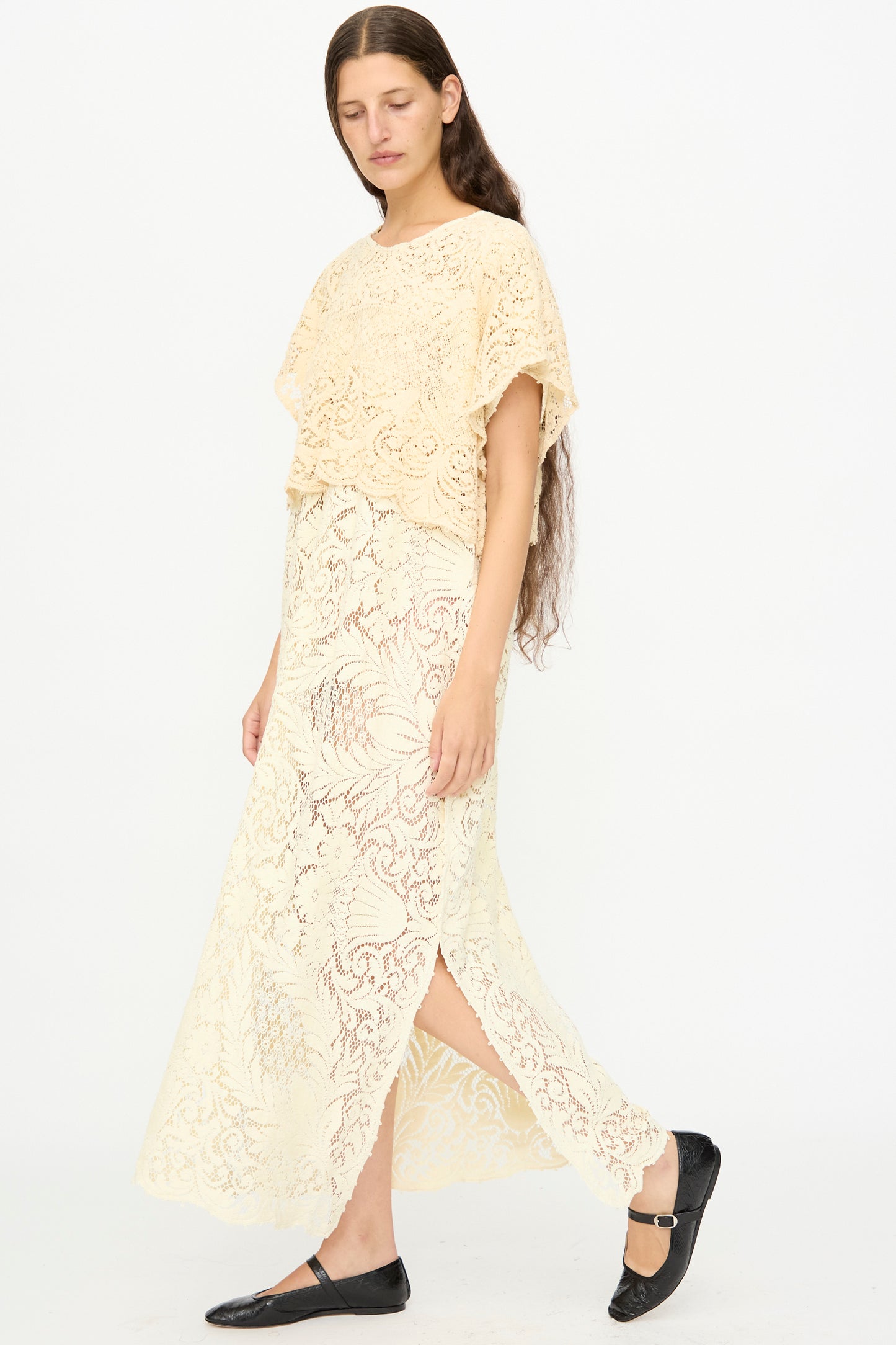 A woman with long brown hair wears the Tilda Gown in Quaker Lace by Thank You Have A Good Day, a cream-hued maxi dress with a side slit, made from antique lace, paired with black flats and set against a plain white background.