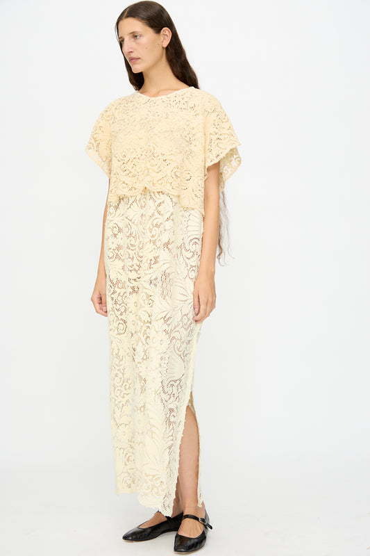 A woman in the Tilda Gown in Quaker Lace by Thank You Have A Good Day, a loose cream maxi dress with floral lace detail and short sleeves, paired with black flats, stands against a plain white background.
