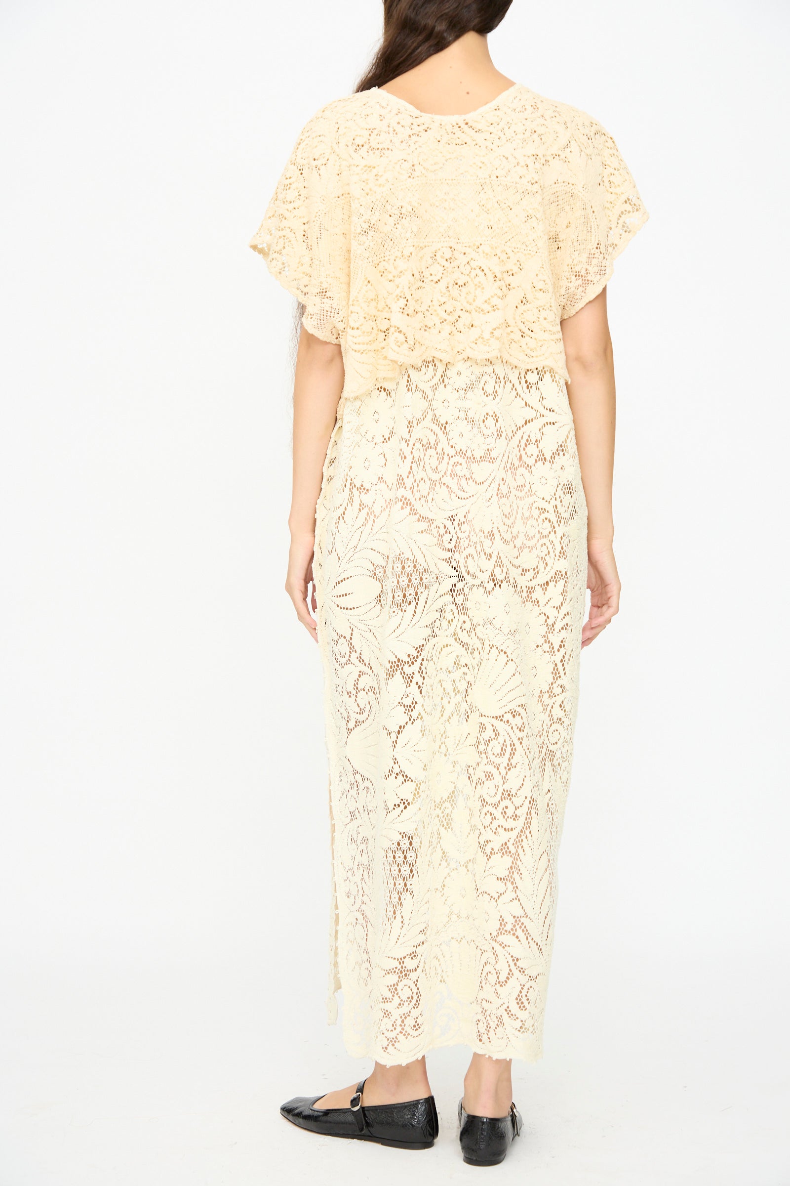 A person wearing the Tilda Gown in Quaker Lace by Thank You Have A Good Day, a cream-colored, floor-length dress, with black flats, seen from the back against a plain white background.