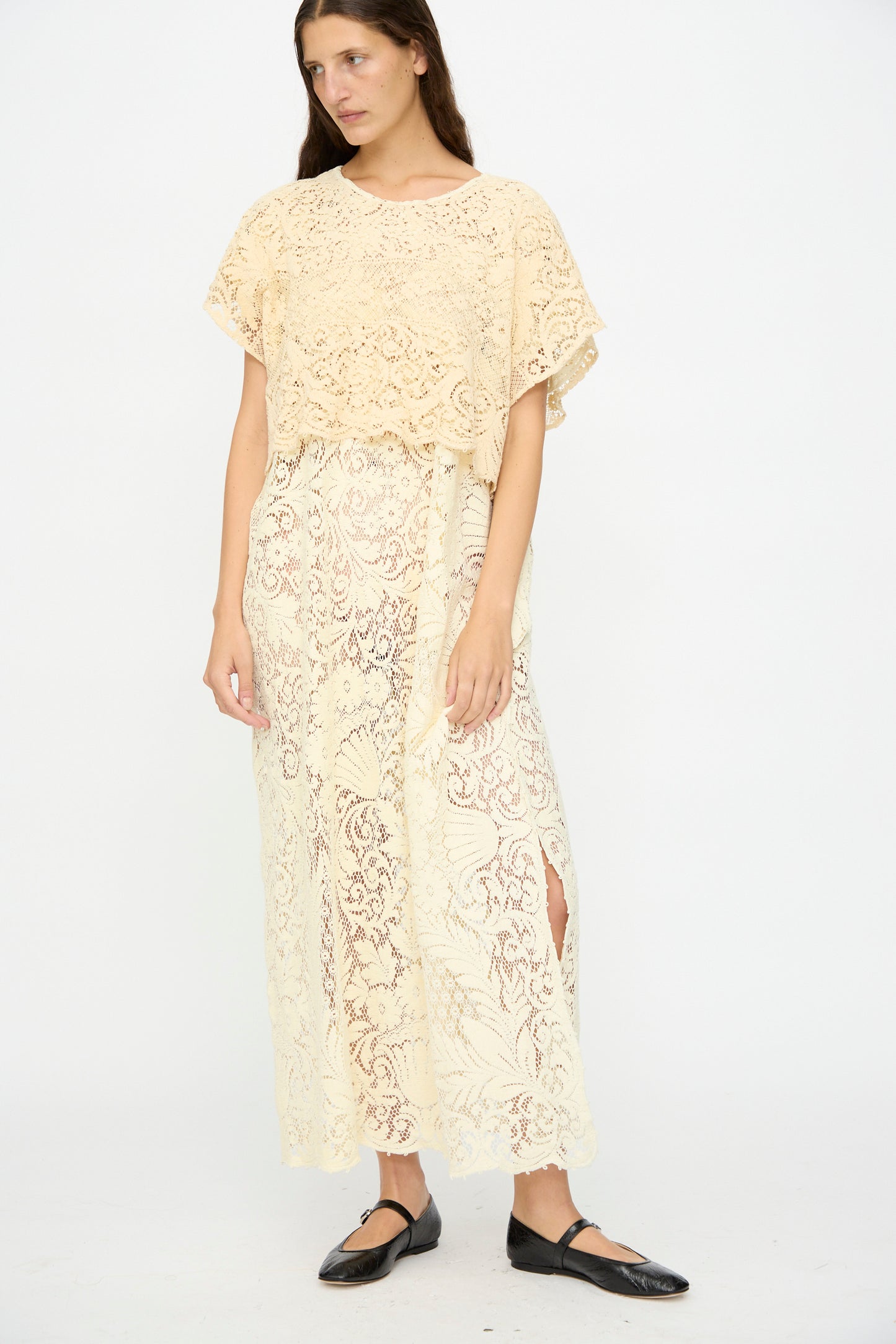 A woman wears the Tilda Gown in Quaker Lace by Thank You Have A Good Day, featuring a loose, cream-colored lace design, paired with black flats, set against a plain white background.
