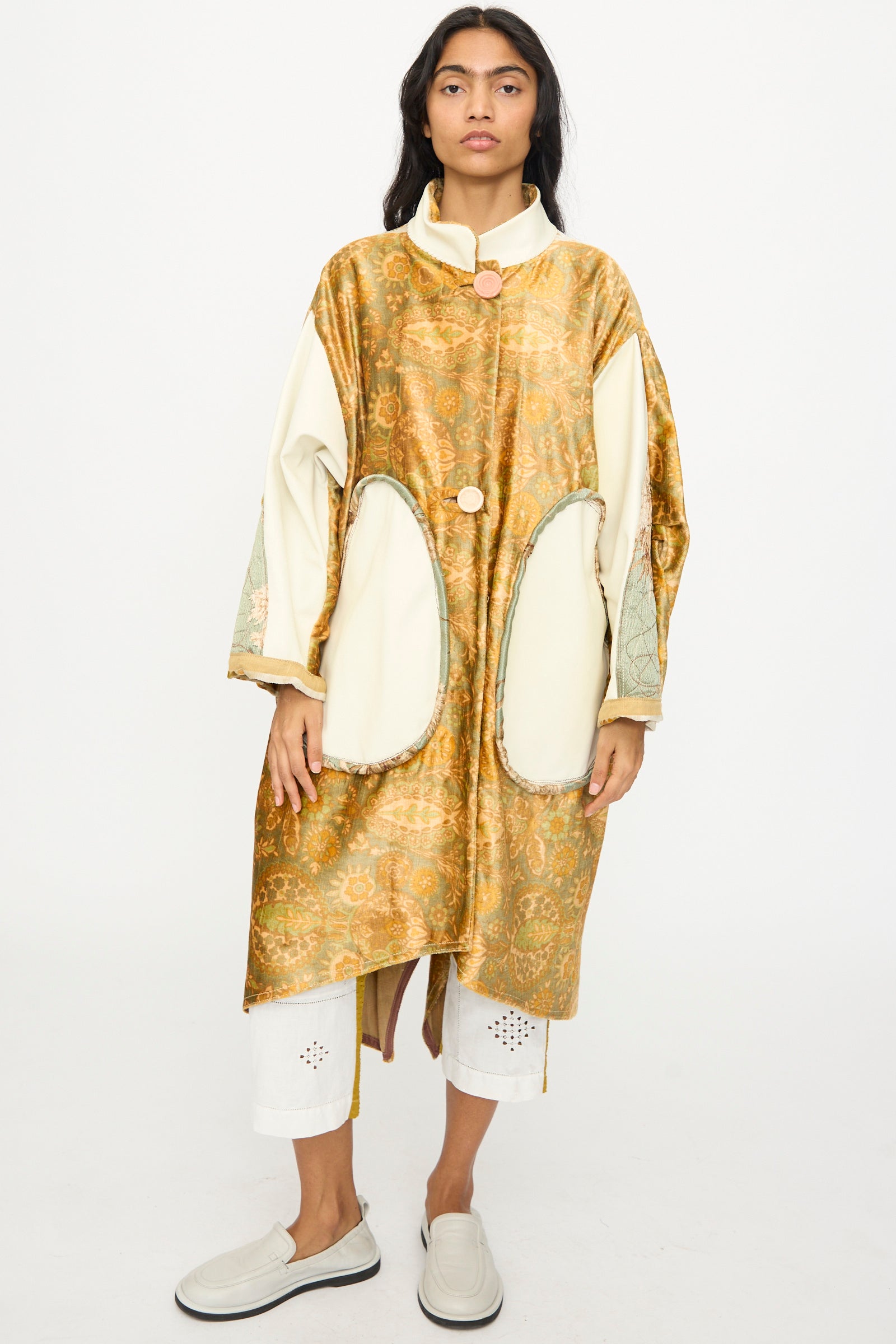 A person stands facing forward, wearing the Velvet and Leather Fishtail Parka by Thank You Have A Good Day in gold and cream, paired with white cropped pants featuring cut-out details and light-colored slip-on shoes.
