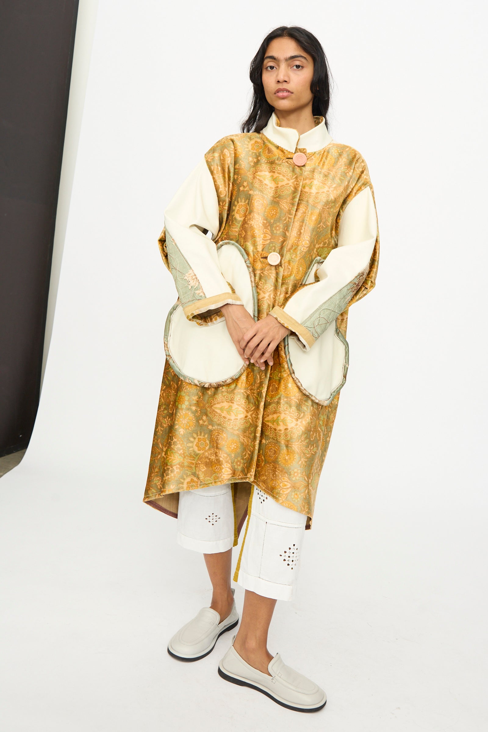 A woman stands against a plain studio backdrop wearing Thank You Have A Good Day’s Velvet and Leather Fishtail Parka, white cropped pants with cut-out details, and light slip-on shoes.