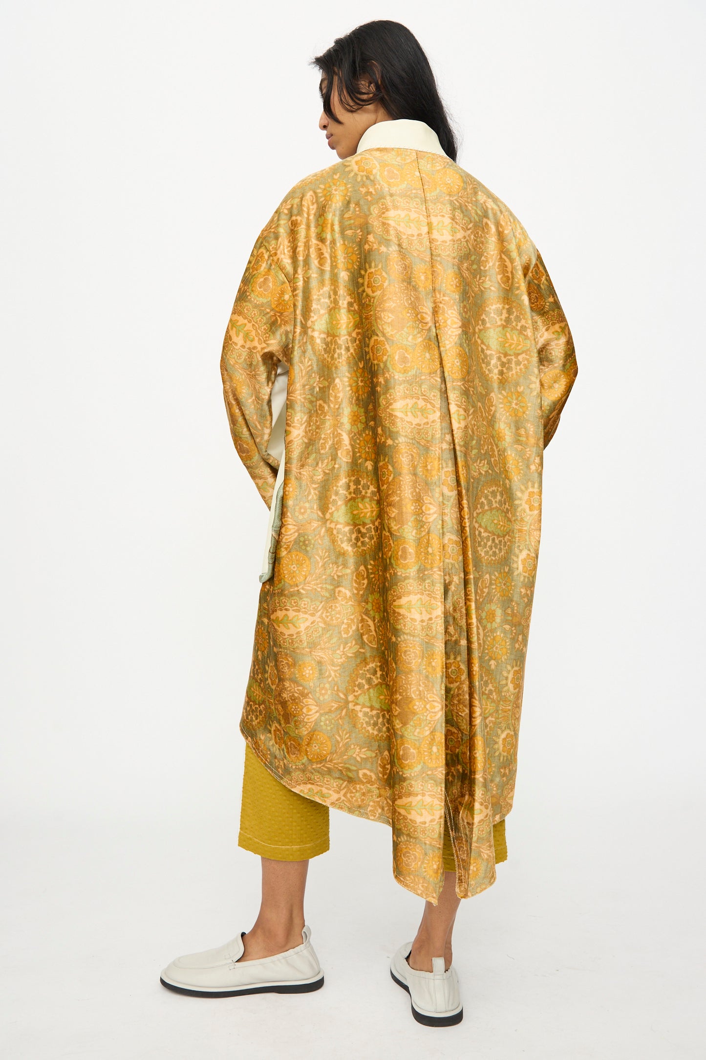 A person stands with back to camera in a gold-patterned Velvet and Leather Fishtail Parka by Thank You Have A Good Day, paired with yellow pants and white slip-on shoes, set against a plain white background.