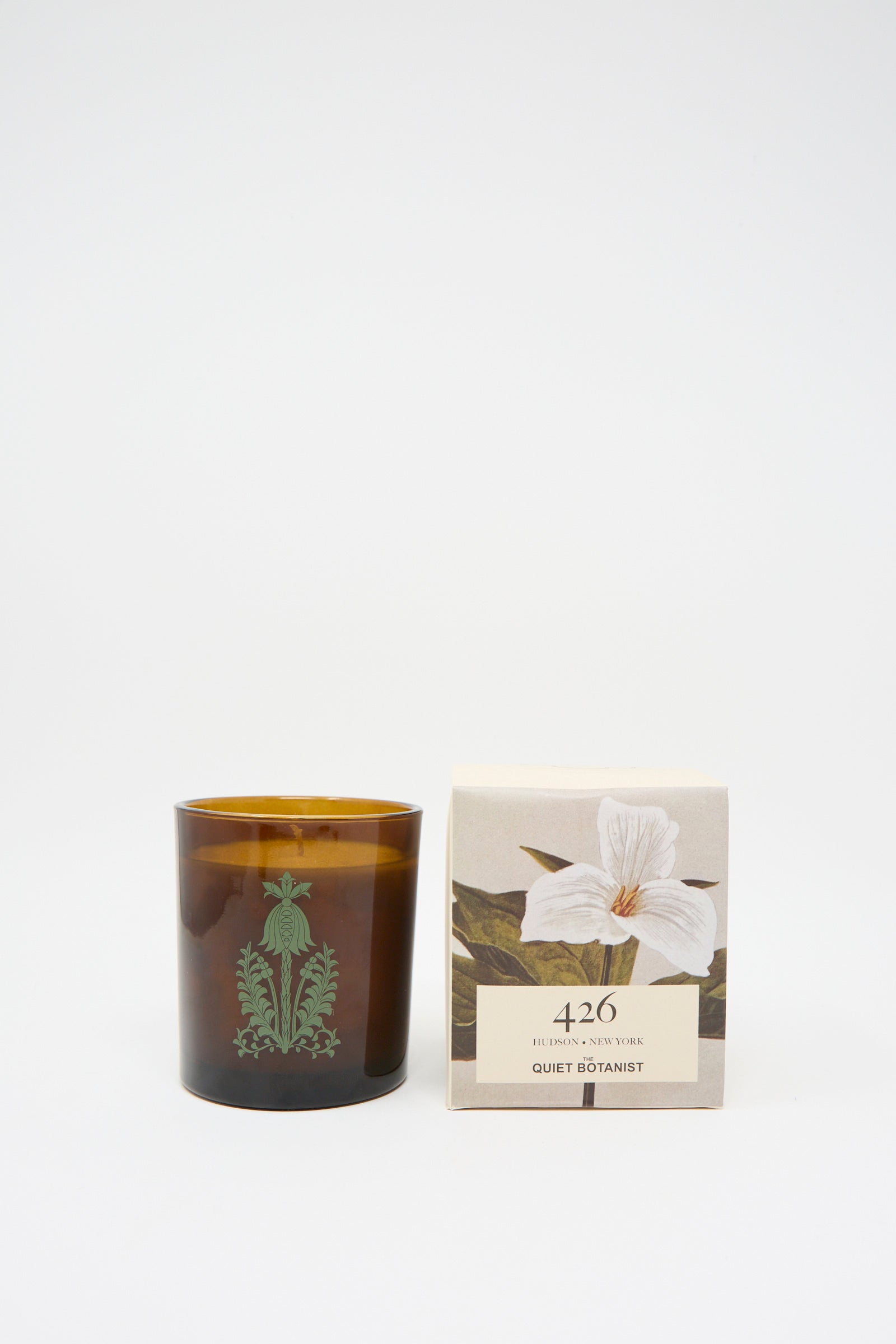 A phthalate-free brown glass candle featuring a plant design is placed next to a beige box decorated with a white flower and the text "The Quiet Botanist 426 Candle," made from natural coconut wax.