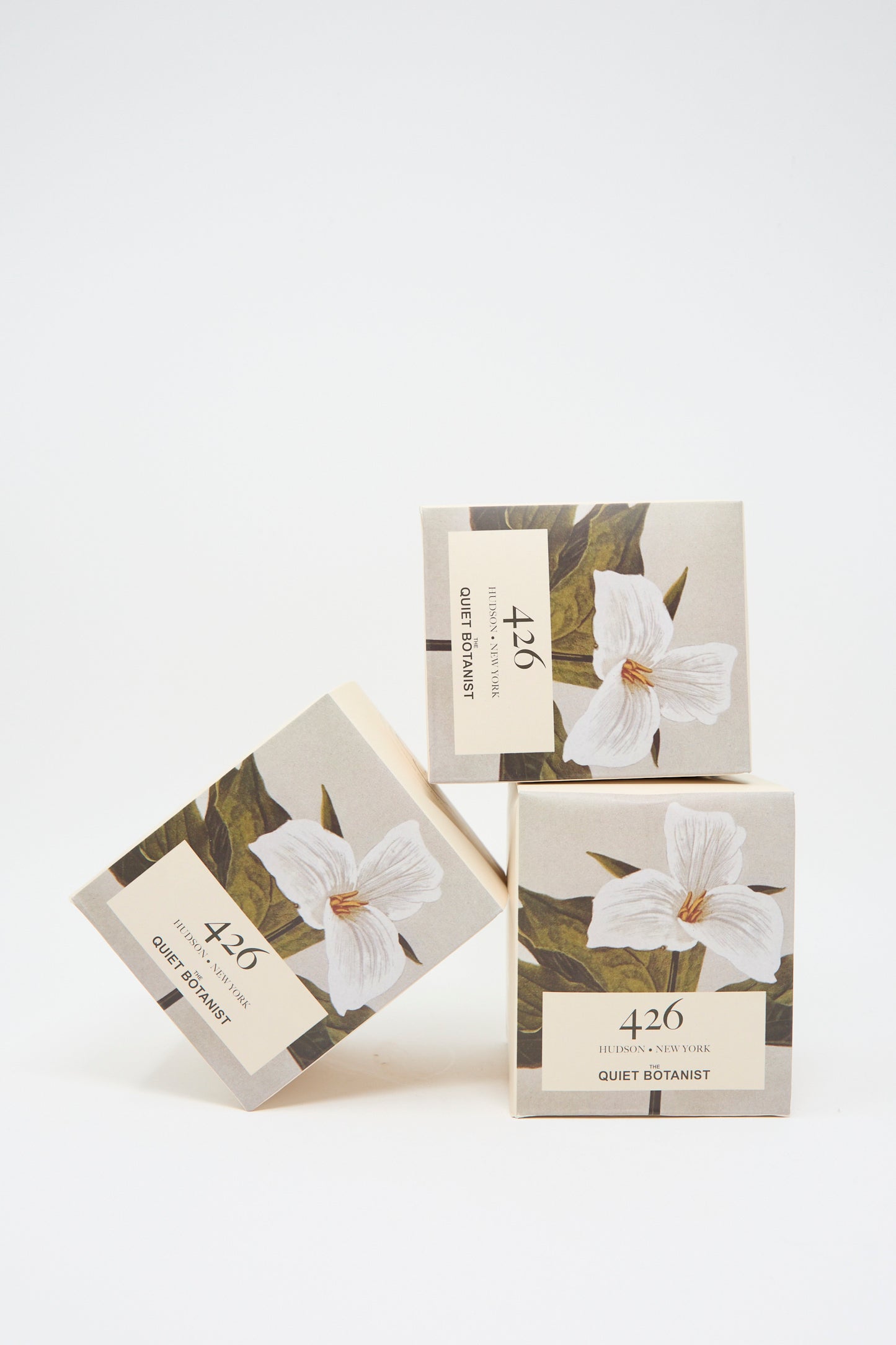 Three stacked boxes featuring floral designs, labeled with "The Quiet Botanist 426 Candle," and a hint of natural coconut wax candle scent against a plain background.