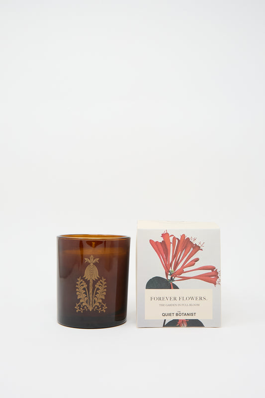 A brown glass candle holder with a botanical design next to a beige box labeled "Forever Flowers Candle" from The Quiet Botanist. The box, featuring an illustration of red flowers, pairs beautifully with a natural beeswax candle.