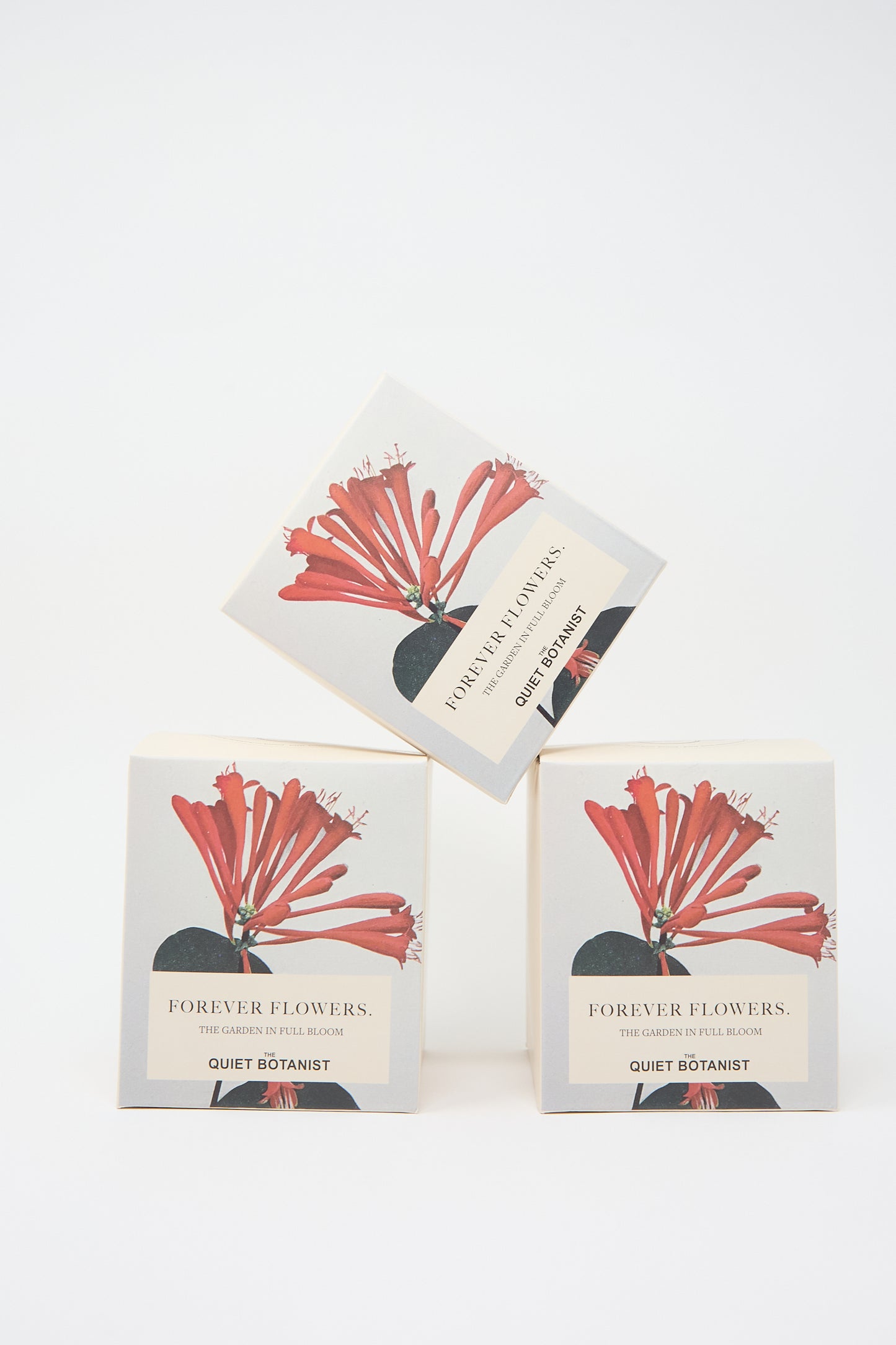 Three boxes labeled "Forever Flowers Candle" from The Quiet Botanist, featuring a red flower illustration. Two boxes are stacked with a third balanced on top. These beautifully designed packages hint at the natural candle inside, crafted from either luxurious coconut wax or pure beeswax.