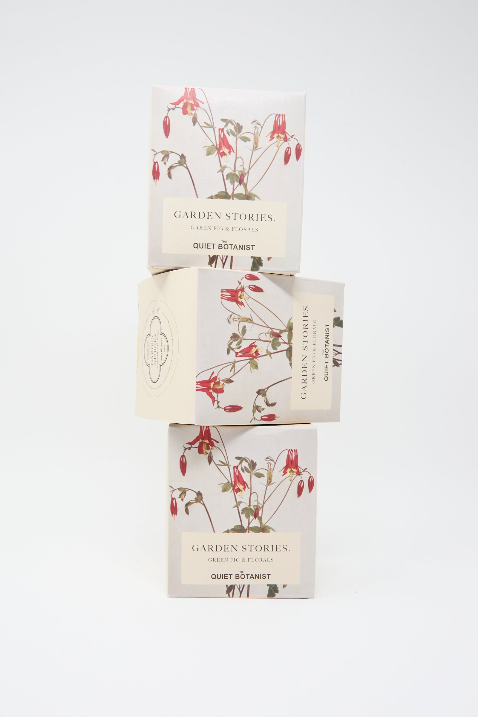 A stack of three boxes labeled "Garden Stories Candle" by The Quiet Botanist, adorned with floral designs, sits elegantly against a plain background; each box contains a natural candle made from coconut wax and free of phthalates.