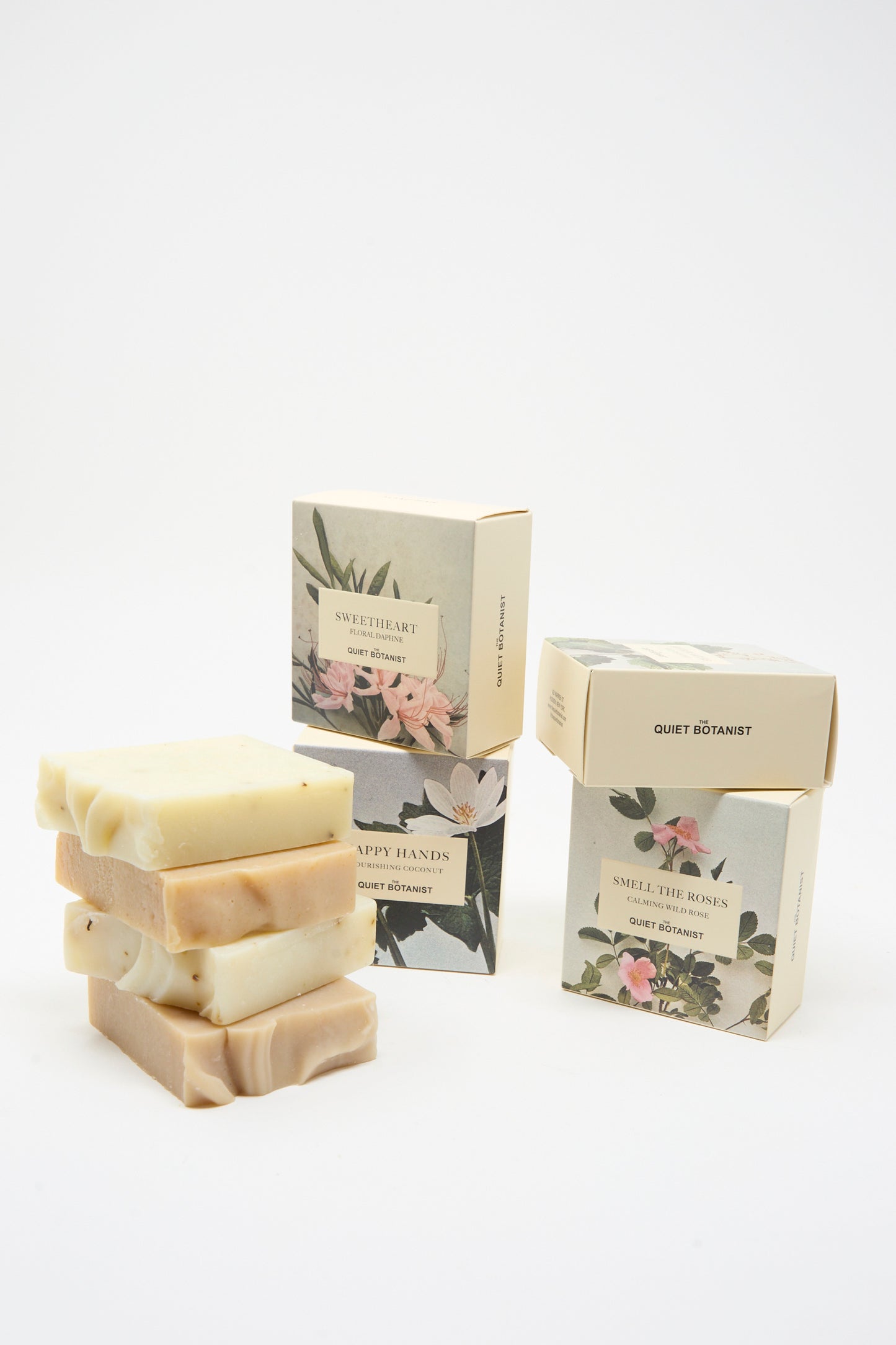 Three bars of The Quiet Botanist Happy Hands Nourishing Coconut Soap are stacked beside three floral-designed boxes displaying the product name, all set against a plain white background.