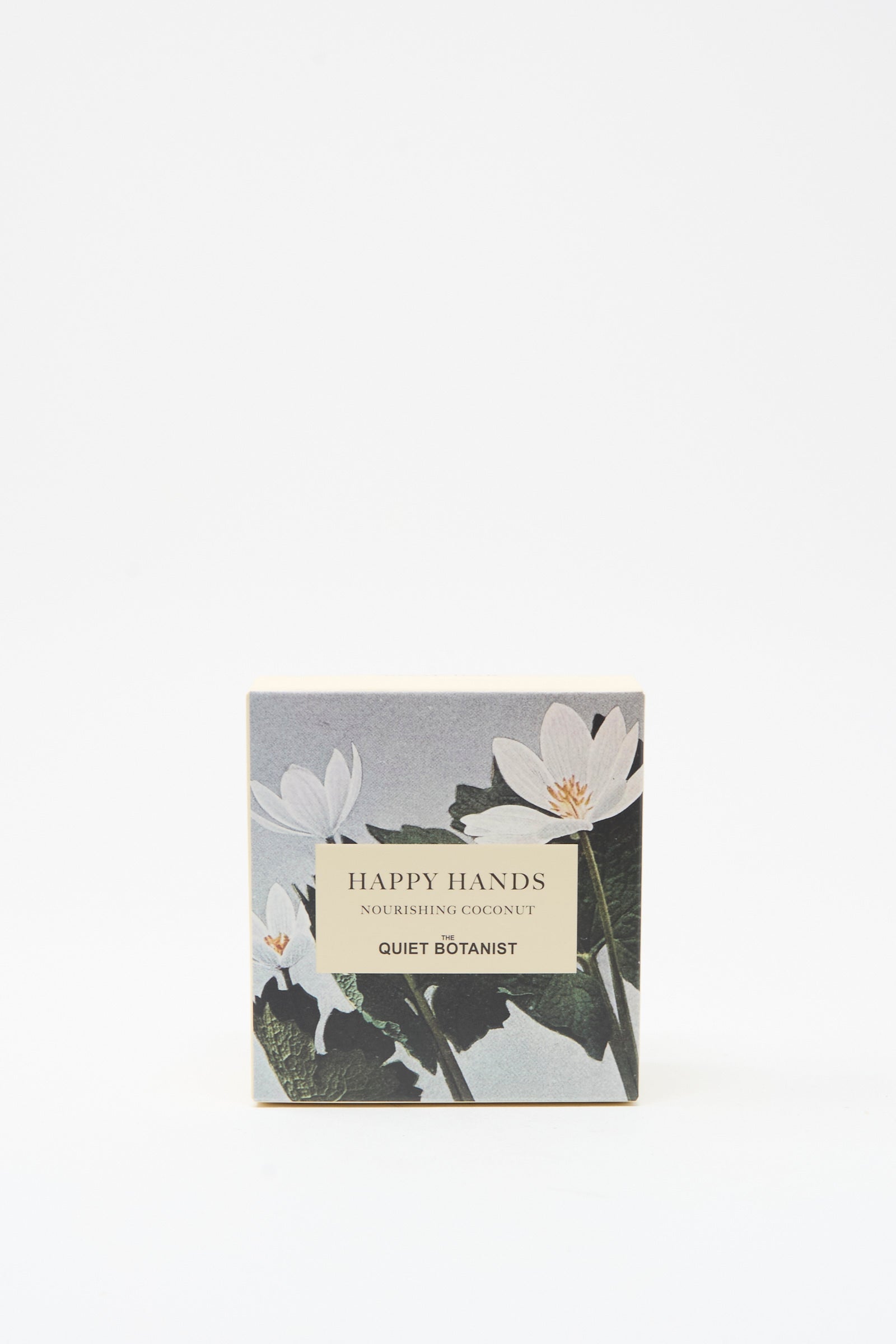 A square box labeled "Happy Hands Nourishing Coconut Soap Bar" by The Quiet Botanist, with white flower designs on a gray background, holding a vegan coconut bar soap handmade in Turkey.