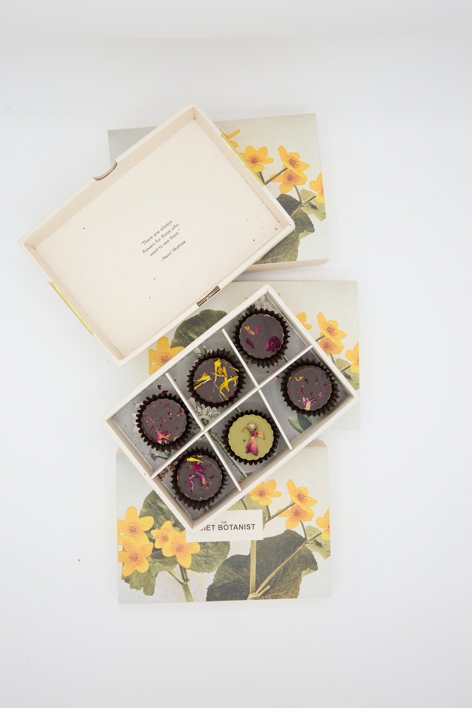 An open box of Holiday Bon Bons by The Quiet Botanist, featuring rose petals and floral designs, rests on a floral-patterned box