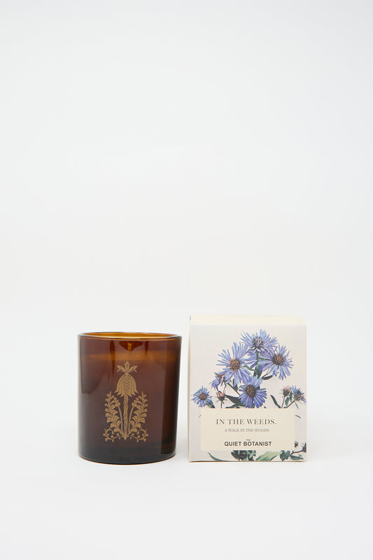 A brown glass candle with a decorative emblem, crafted from natural coconut wax, sits next to a box labeled "In the Weeds Candle" by The Quiet Botanist, adorned with a floral design on a plain white background.