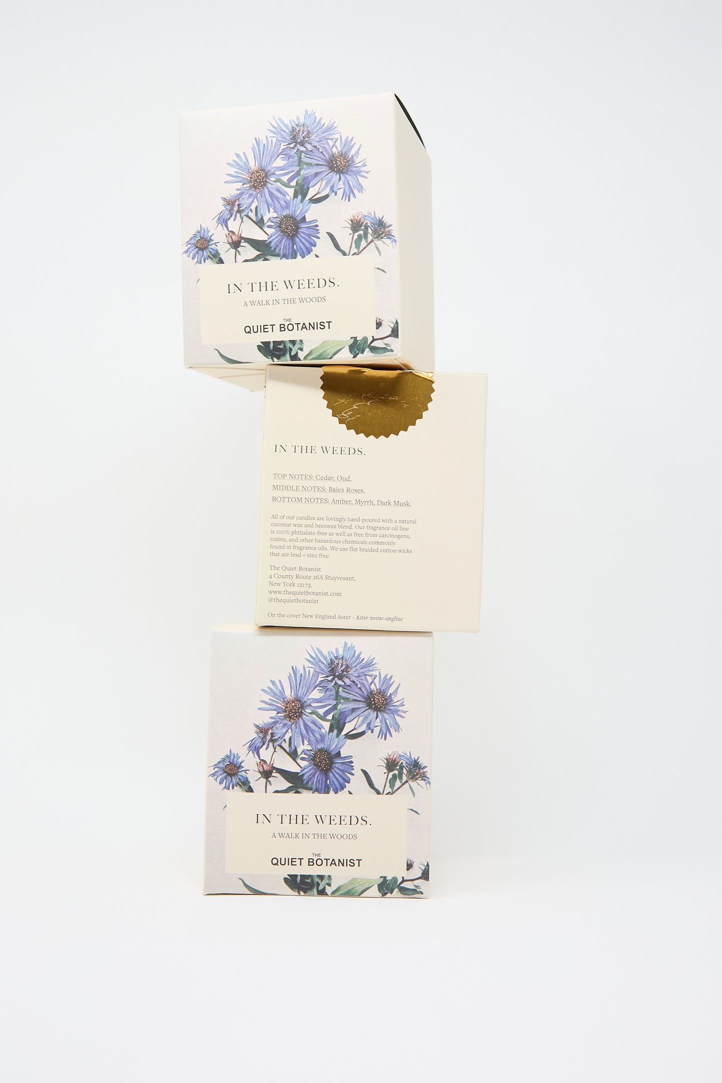 Three stacked boxes of In the Weeds Candle by The Quiet Botanist. Made with natural coconut wax, the packaging features botanical illustrations and a gold seal.