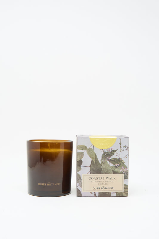 A brown glass candle labeled “The Quiet Botanist” and made from natural coconut wax is displayed beside a box featuring botanical illustrations and the name “Limited Edition Summer Candle Coastal Walk,” all set against a plain white background.
