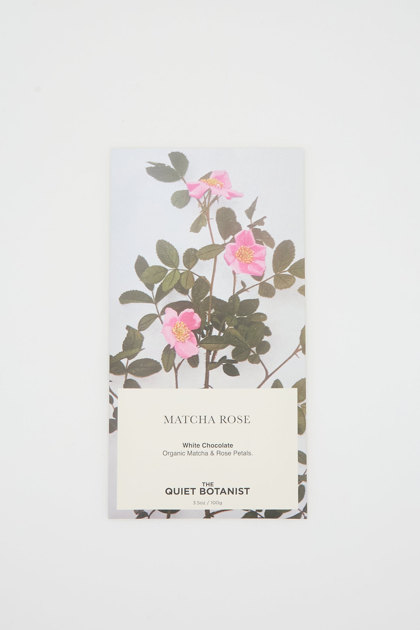 A Matcha Rose Chocolate Bar from The Quiet Botanist, featuring packaging adorned with pink flowers and green leaves on a white background. The text reveals it is white chocolate infused with organic matcha and rose petals.