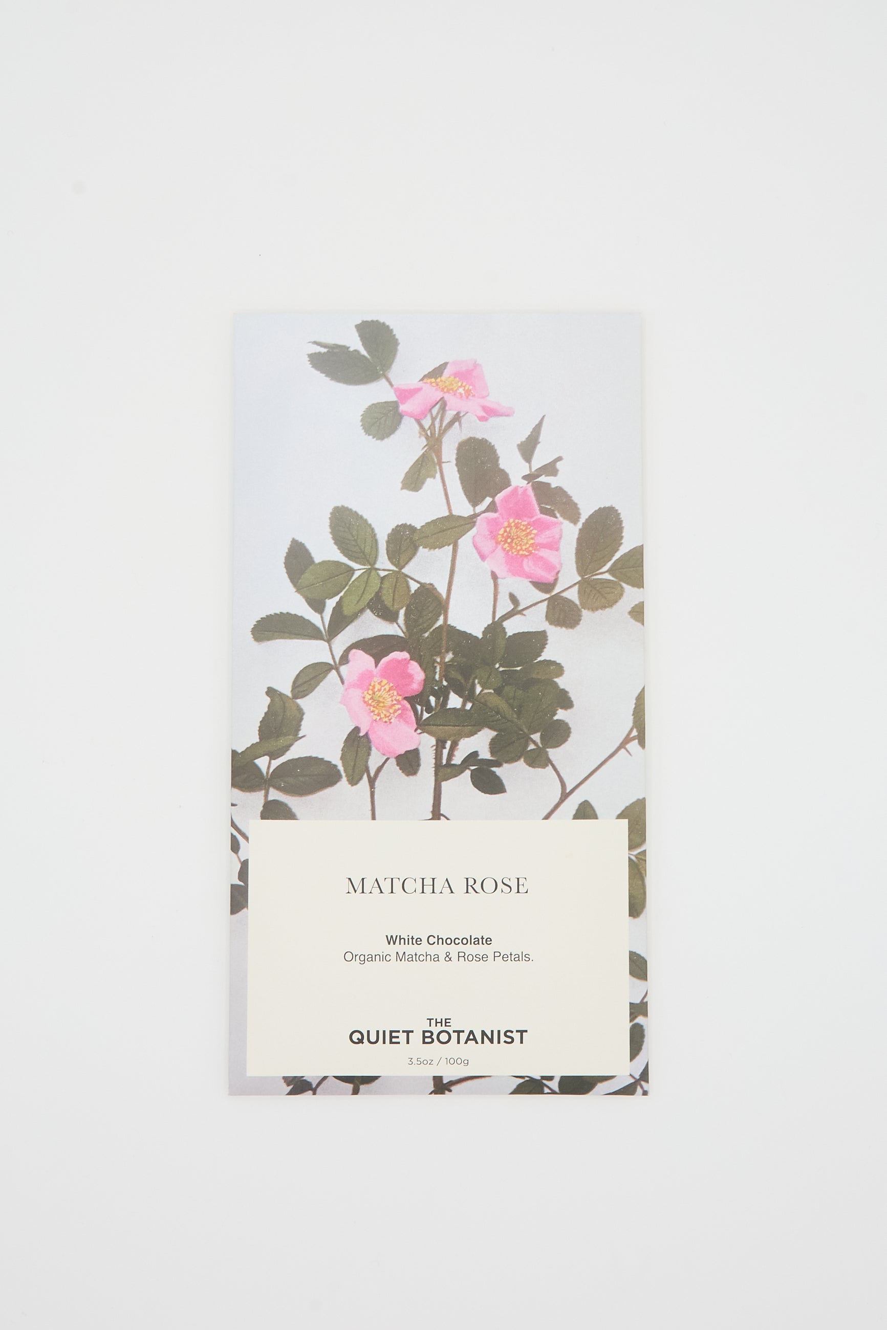 A Matcha Rose Chocolate Bar from The Quiet Botanist, featuring packaging adorned with pink flowers and green leaves on a white background. The text reveals it is white chocolate infused with organic matcha and rose petals.