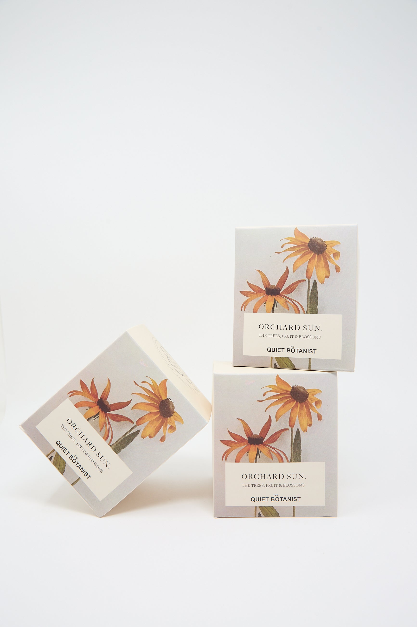 Three boxes of "Orchard Sun Candle" by The Quiet Botanist are displayed, with two boxes stacked and one box tilted on the side. The design features yellow-orange flowers on a light grey background, reflecting the natural charm of these beeswax candles.