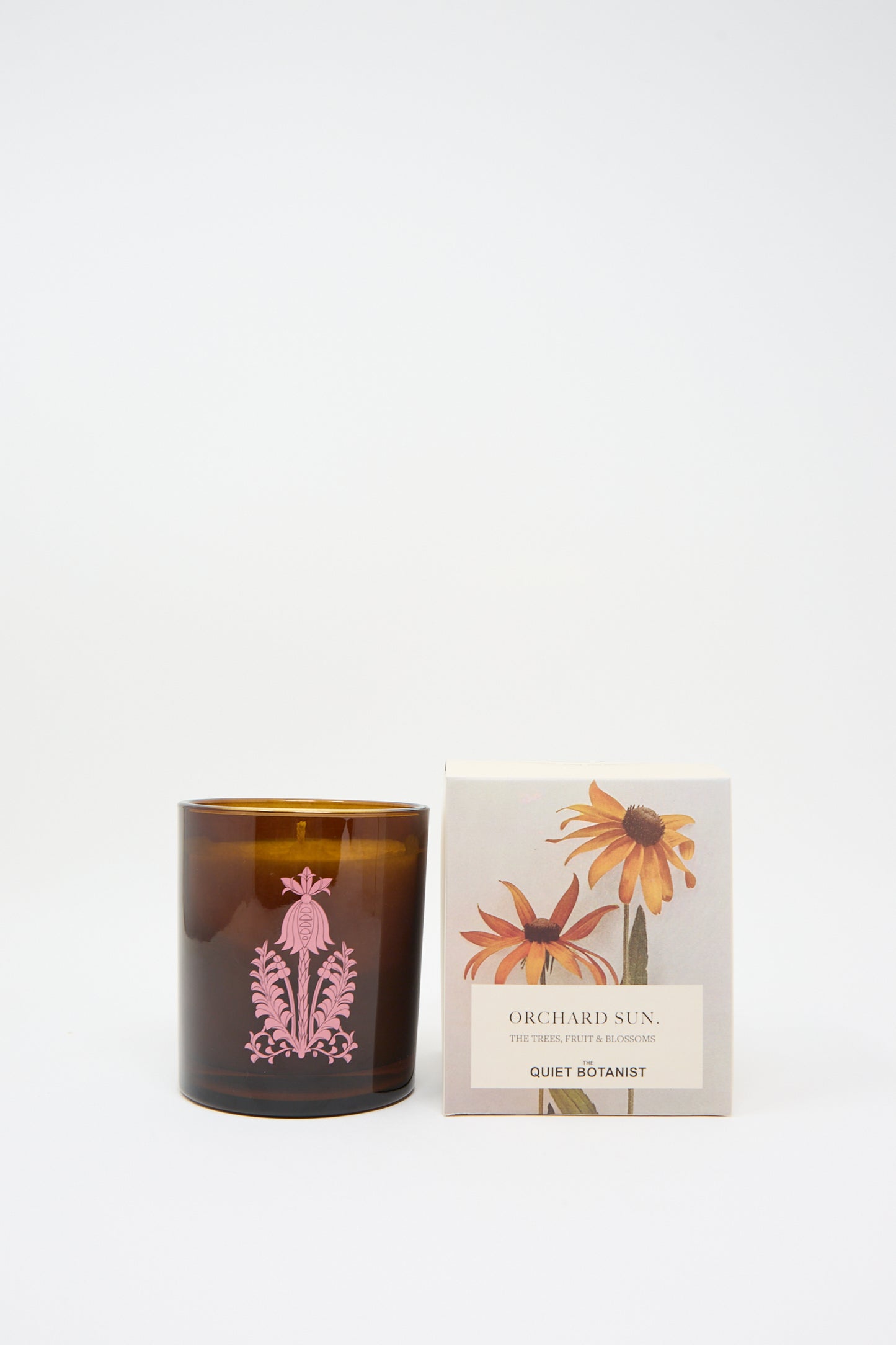 A brown glass candle holder with a pink floral design sits next to a box labeled "Orchard Sun Candle" by The Quiet Botanist, featuring yellow and orange flowers. This elegant set includes a natural candle made from coconut wax.