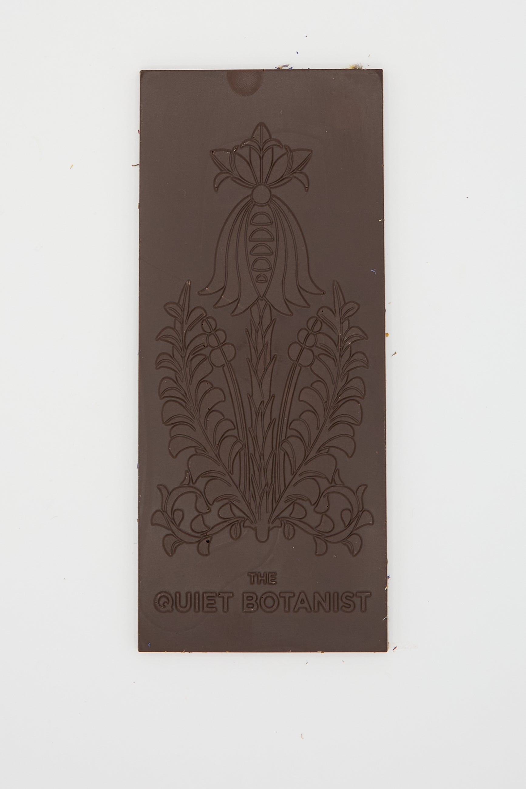 A rectangular piece of organic dark chocolate adorned with an embossed floral design and "The Quiet Botanist" at the bottom. This delightful treat, called the Wildflower Dream Bar, encapsulates the essence of nature's dried wildflowers in every bite.