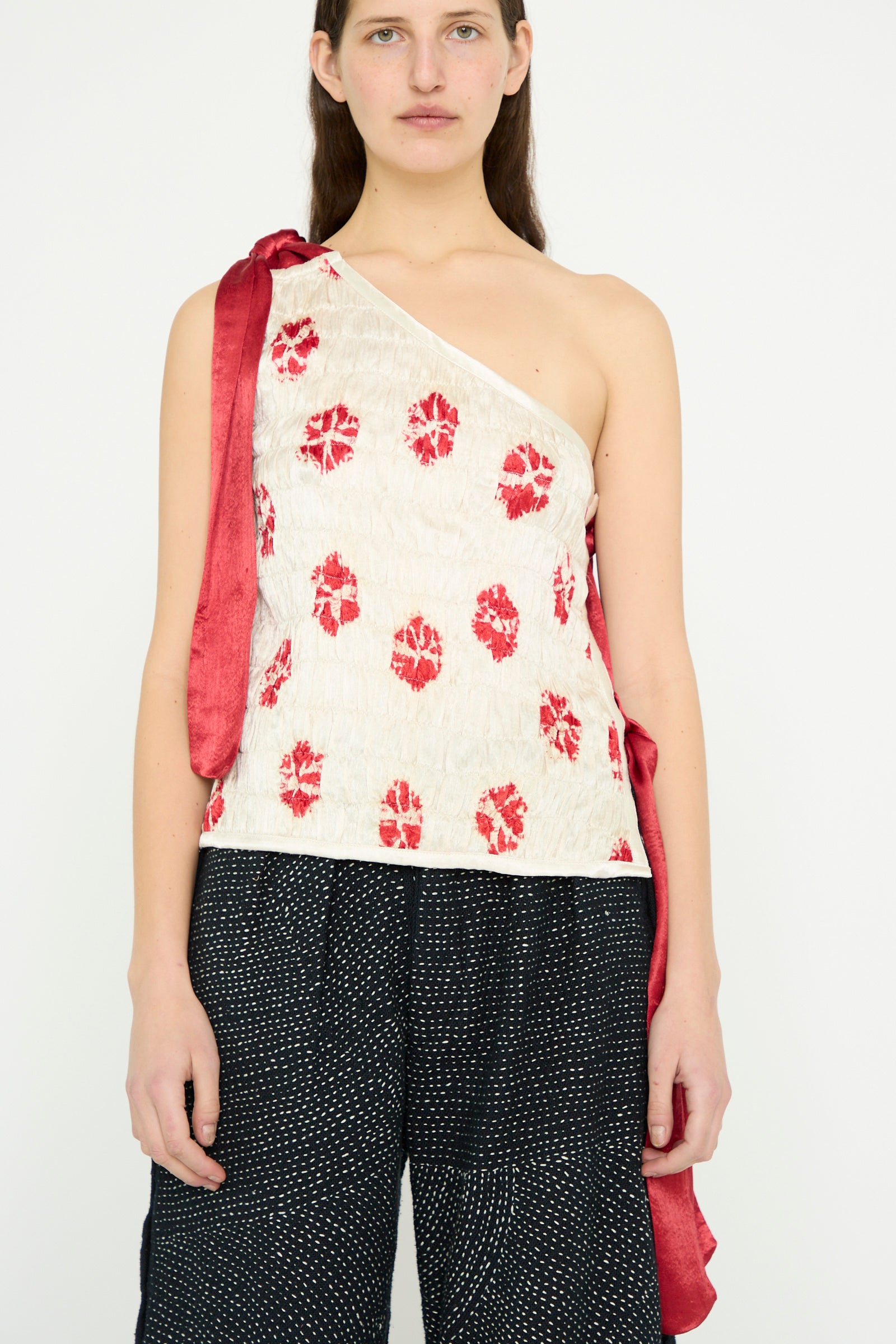 A model wears the Gathered Mashroo Knot Tie Top Bandhej Flowers in White with Red by TIGRA TIGRA, featuring a one-shoulder design with red floral patterns and a red strap, paired with dark patterned pants.