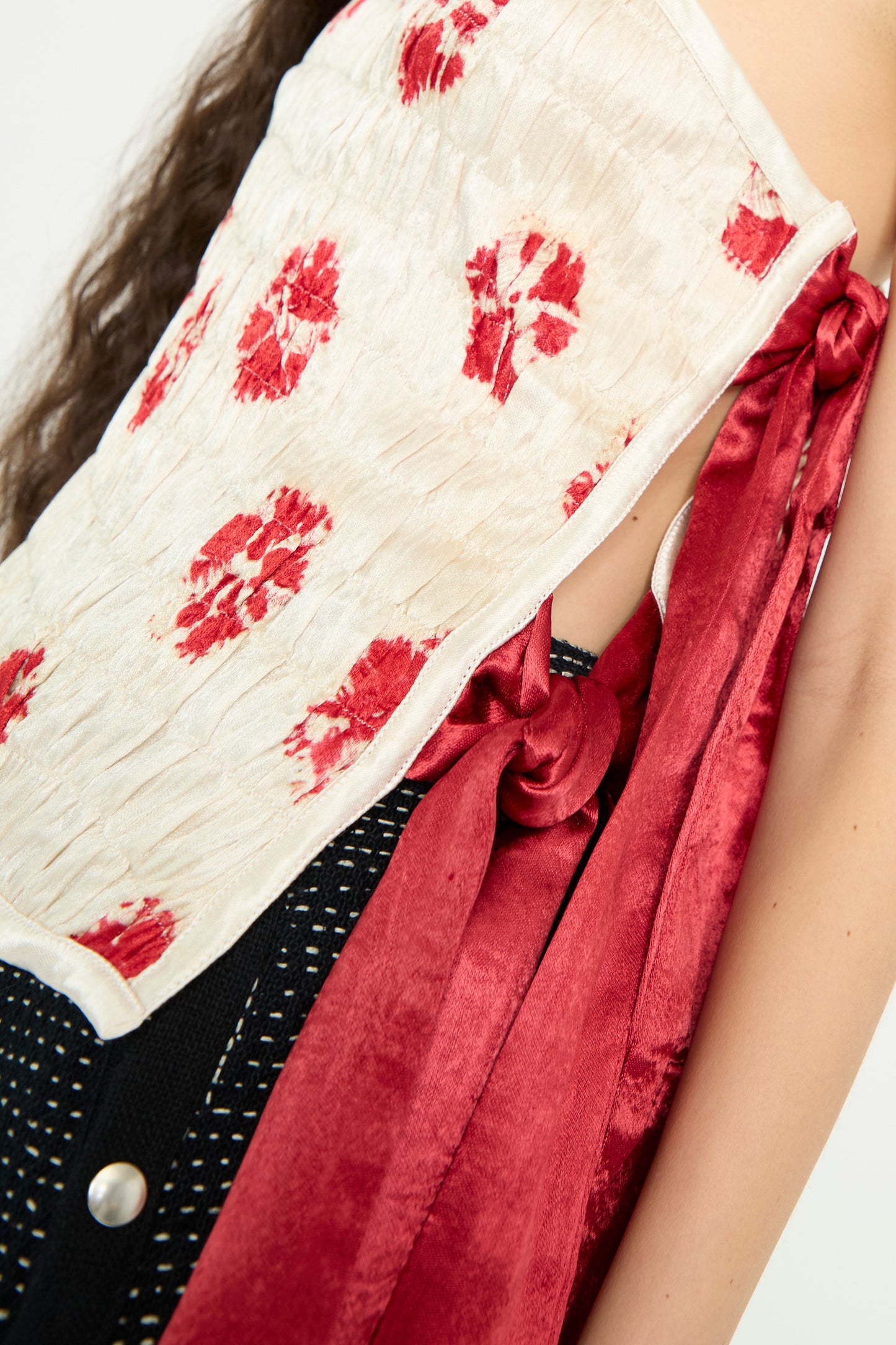 A close-up of a model's torso, wearing the TIGRA TIGRA Gathered Mashroo Knot Tie Top Bandhej Flowers in White with Red, against a plain backdrop. 