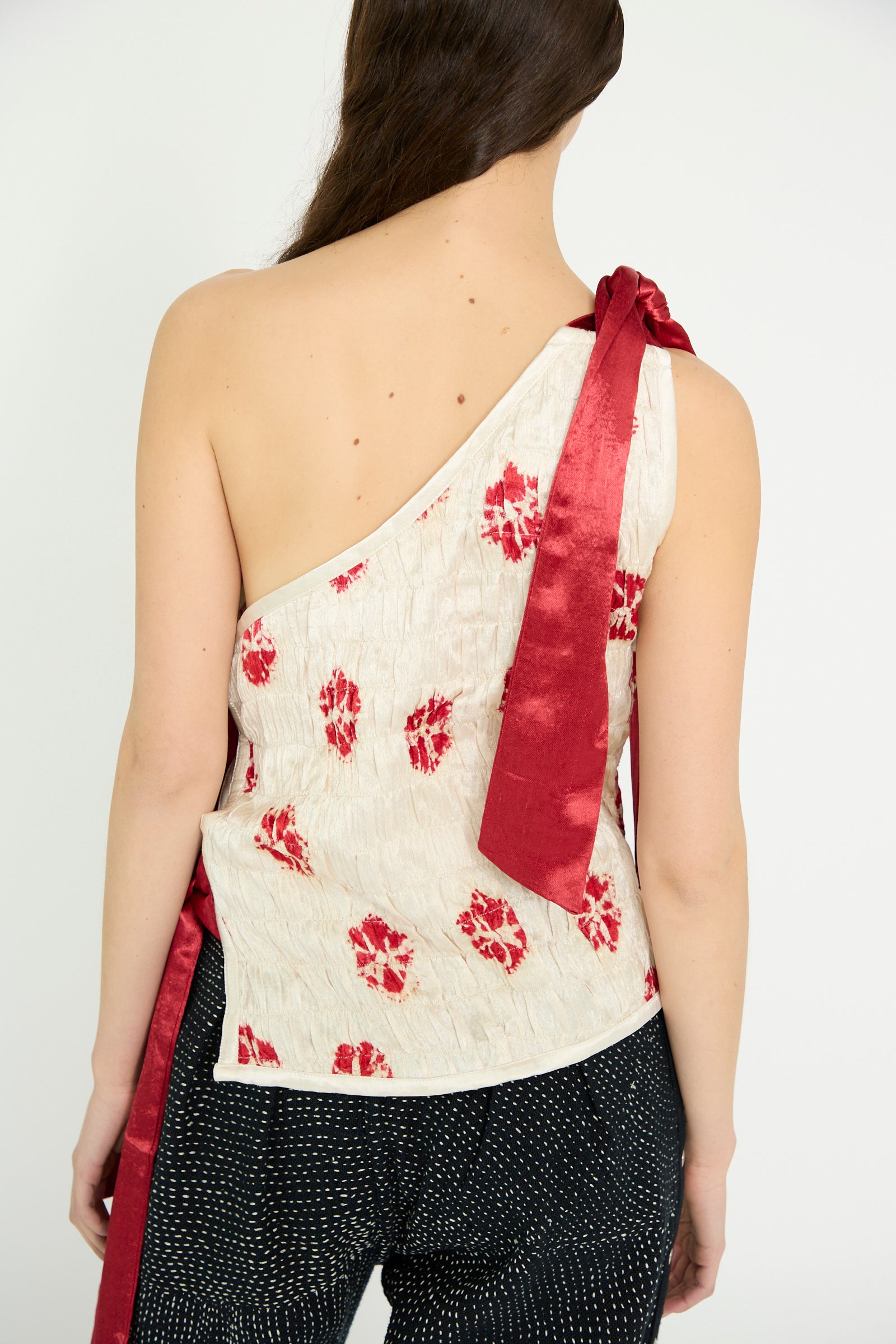 A model is seen from behind wearing the TIGRA TIGRA Gathered Mashroo Knot Tie Top Bandhej Flowers in White with Red, paired with black pants featuring white dots, against a white backdrop. 