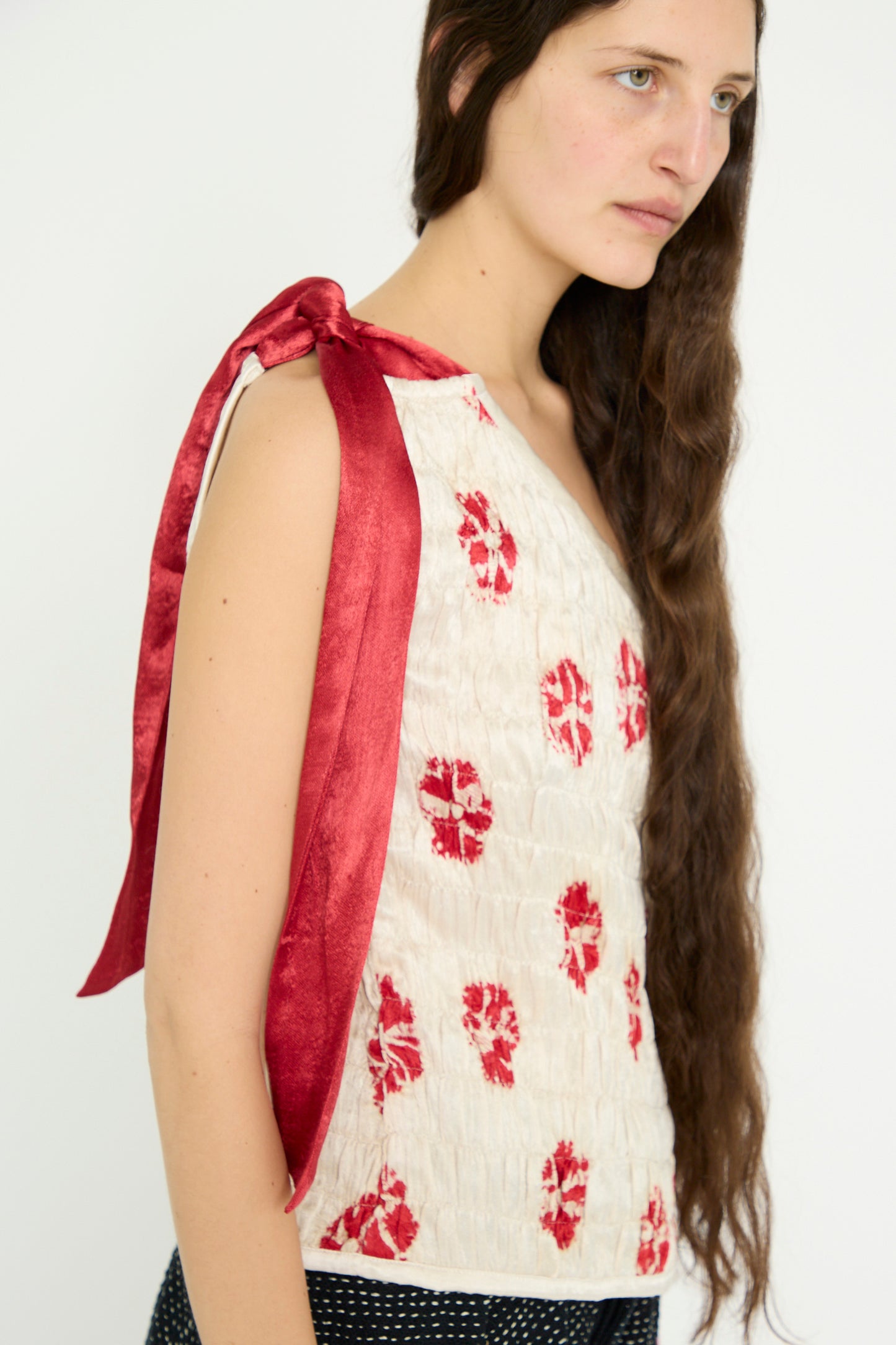 A model with long hair is wearing a TIGRA TIGRA Gathered Mashroo Knot Tie Top featuring Bandhej Flowers in White with Red and a red bow on the shoulder, paired with black pants, against a white background. 