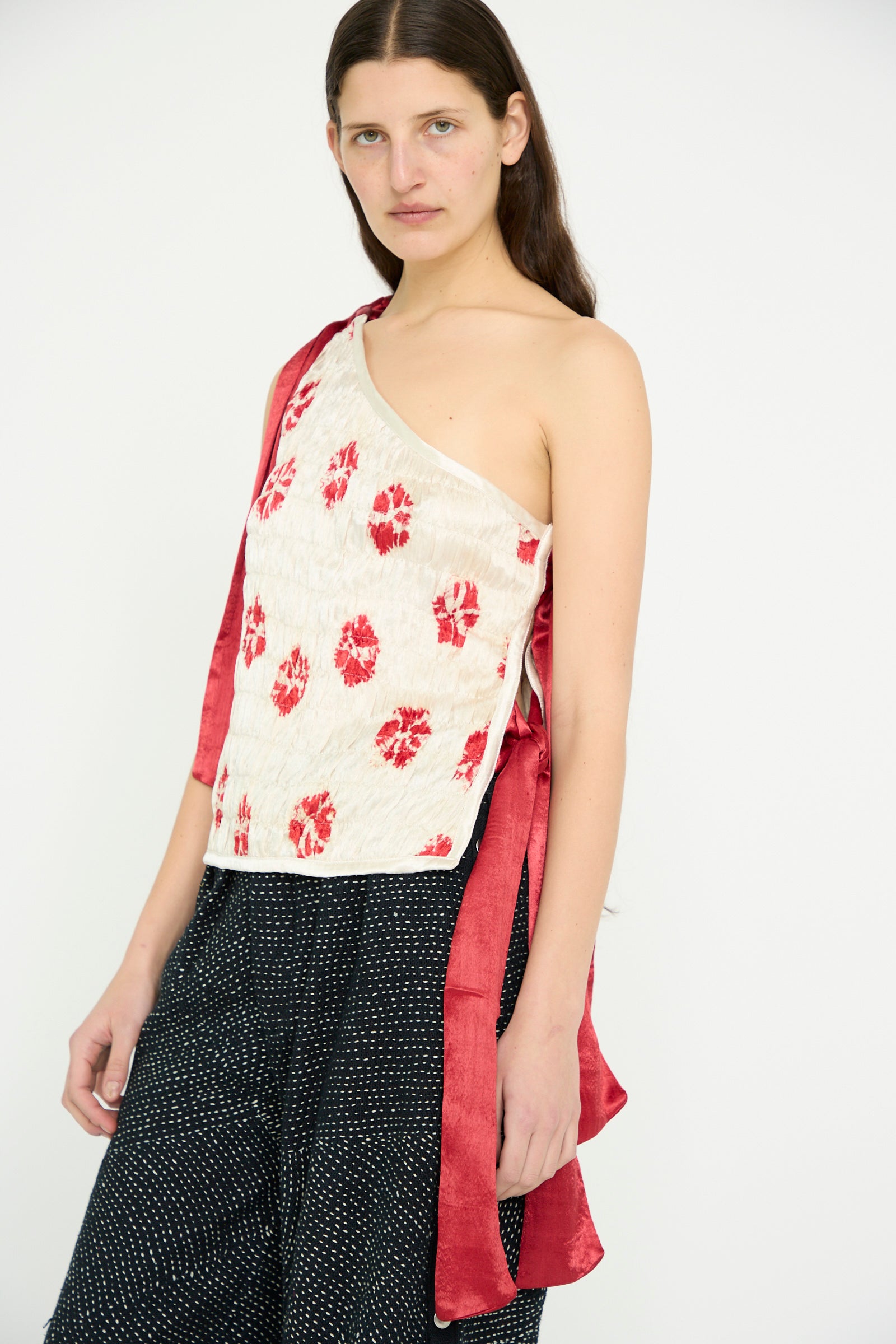 A model wears the TIGRA TIGRA Gathered Mashroo Knot Tie Top featuring red Bandhej flowers on white, paired with a black dotted pant and a red Mashroo Silk fabric draped over their shoulder.