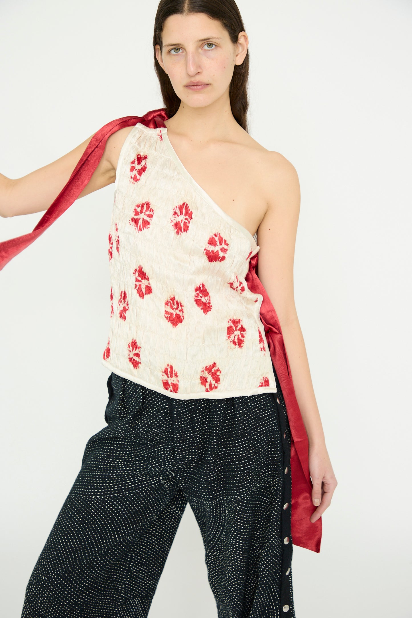 A person stands against a plain backdrop, showcasing the TIGRA TIGRA Gathered Mashroo Knot Tie Top Bandhej Flowers in White with Red, paired with dark pants.