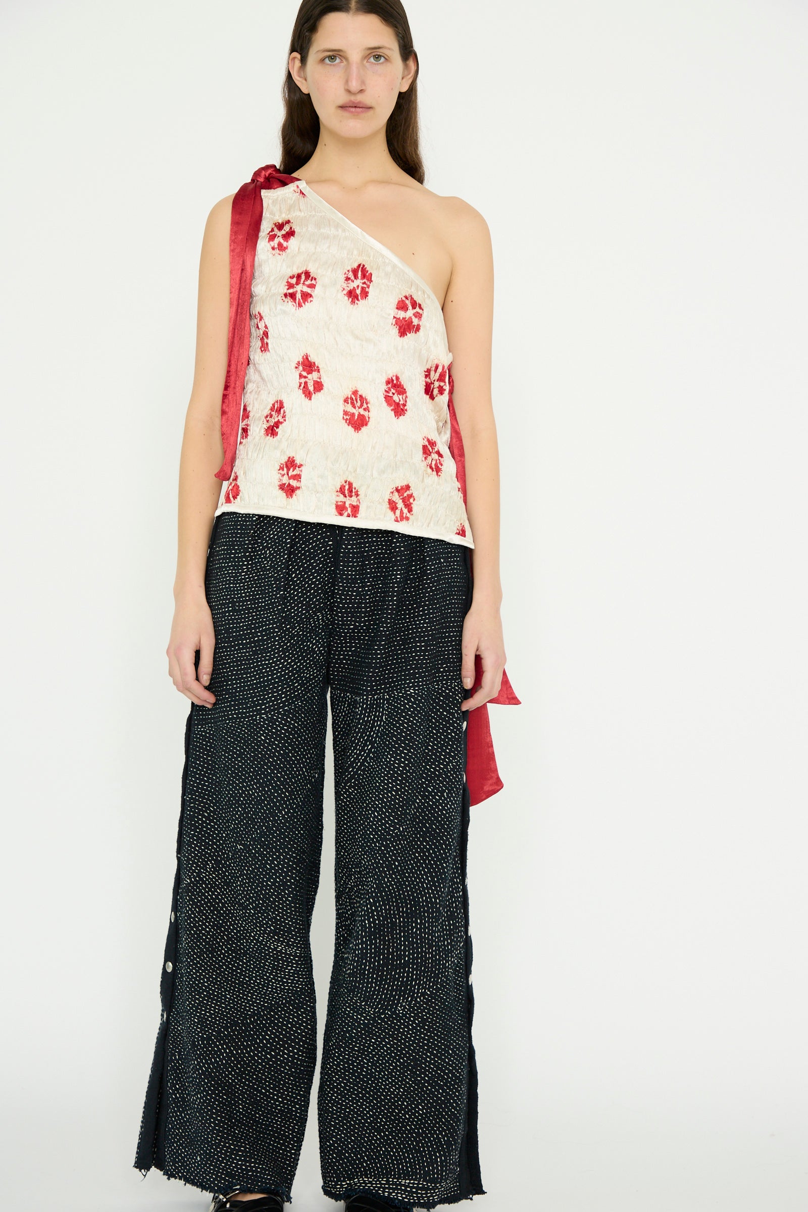 A person is wearing a one-shoulder top featuring a red and white pattern, paired with TIGRA TIGRA's Heavy Khadi Kantha Snap Pant in Black with Cream. They stand against a plain background.