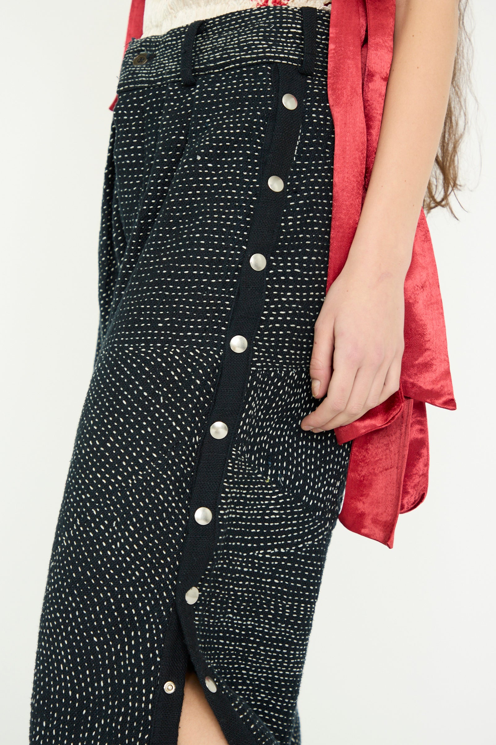 A close-up of a person wearing the Heavy Khadi Kantha Snap Pant in Black with Cream from TIGRA TIGRA, paired with a red top, arm on their side. 