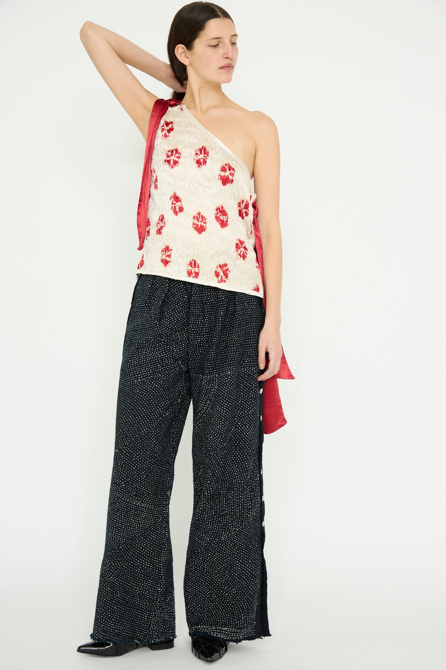 Against a white backdrop, a model is seen in TIGRA TIGRA's one-shoulder top featuring red florals paired with Heavy Khadi Kantha Snap Pant in Black with Cream, with one arm raised.