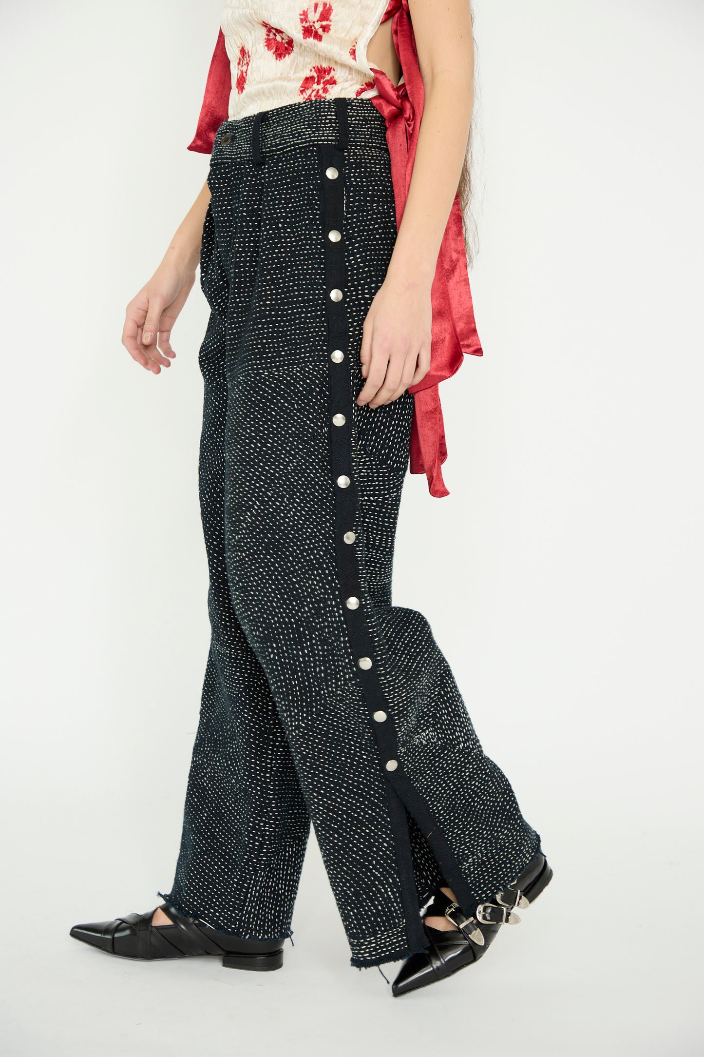 A person walks against a white backdrop, A model facing the side wearing TIGRA TIGRA Heavy Khadi Kantha Snap Pants in Black with Cream, featuring side buttons, against a plain background. 