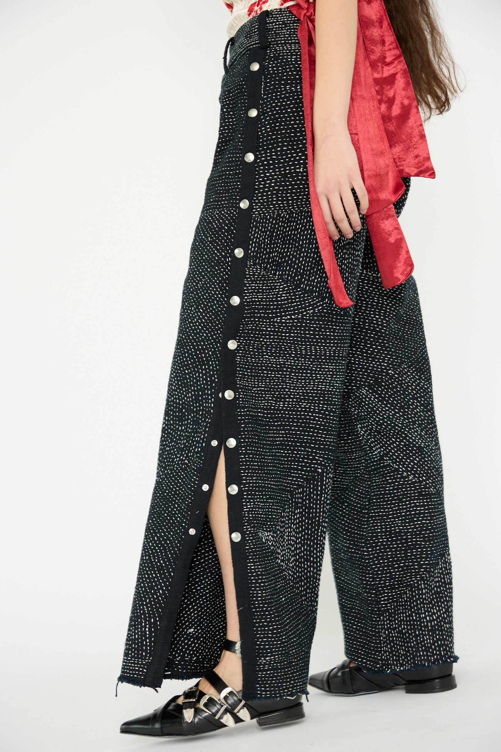 A model wearing the TIGRA TIGRA Heavy Khadi Kantha Snap Pant in Black with Cream, featuring a handwoven texture and side buttons. Paired with a red top and black sandals against a white background. 