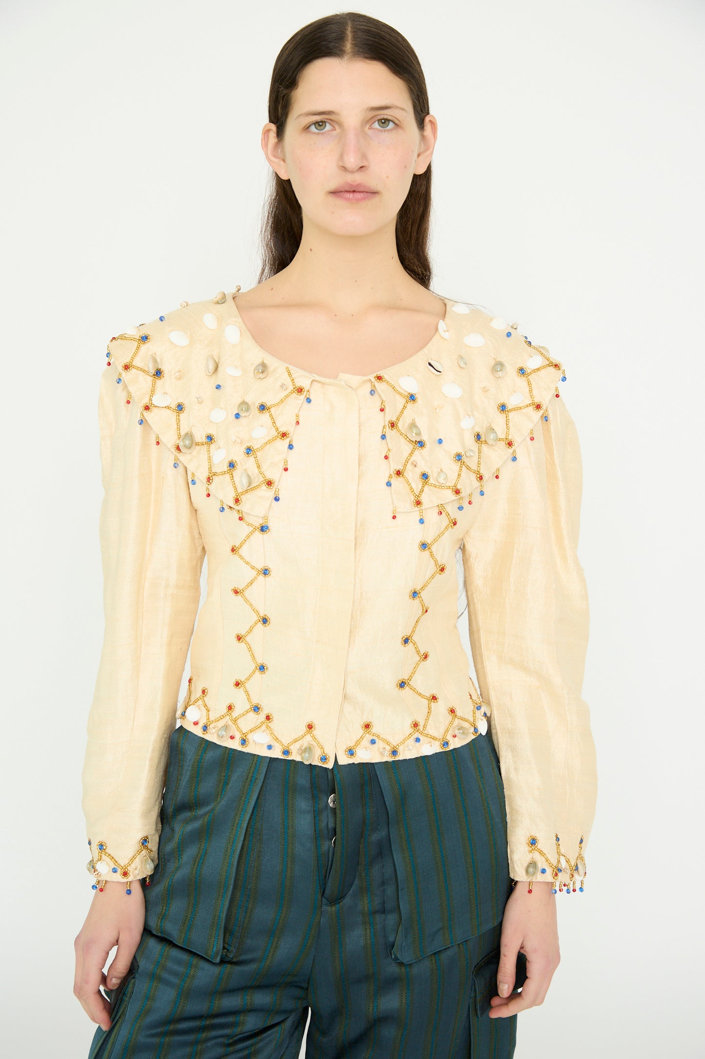 A model wears the TIGRA TIGRA Raw Silk Pirate Top with Cowrie Shells in cream and gold, paired with teal striped pants, standing against a plain background.