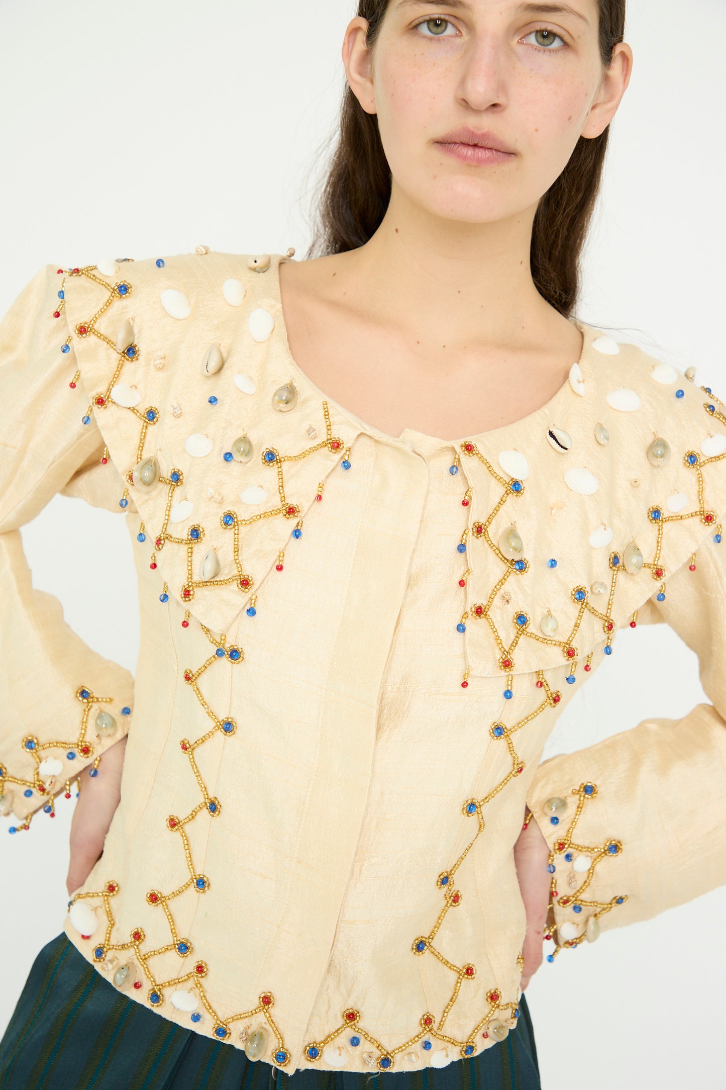 A model stands with hands on their hips against a plain background wearing the Raw Silk Pirate Top with Cowrie Shells in Cream with Gold by TIGRA TIGRA, featuring geometric bead embellishments and a scoop-neck resembling raw silk.