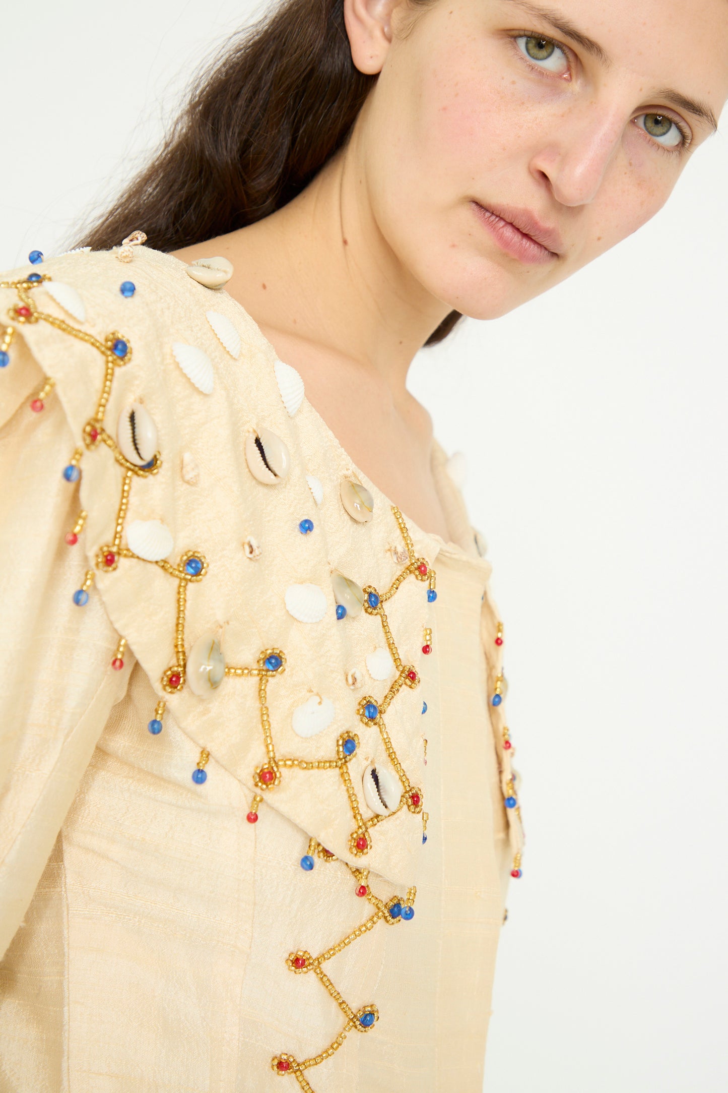 A model wearing the Raw Silk Pirate Top with Cowrie Shells in Cream with Gold by TIGRA TIGRA gazes at the camera against a plain white background.