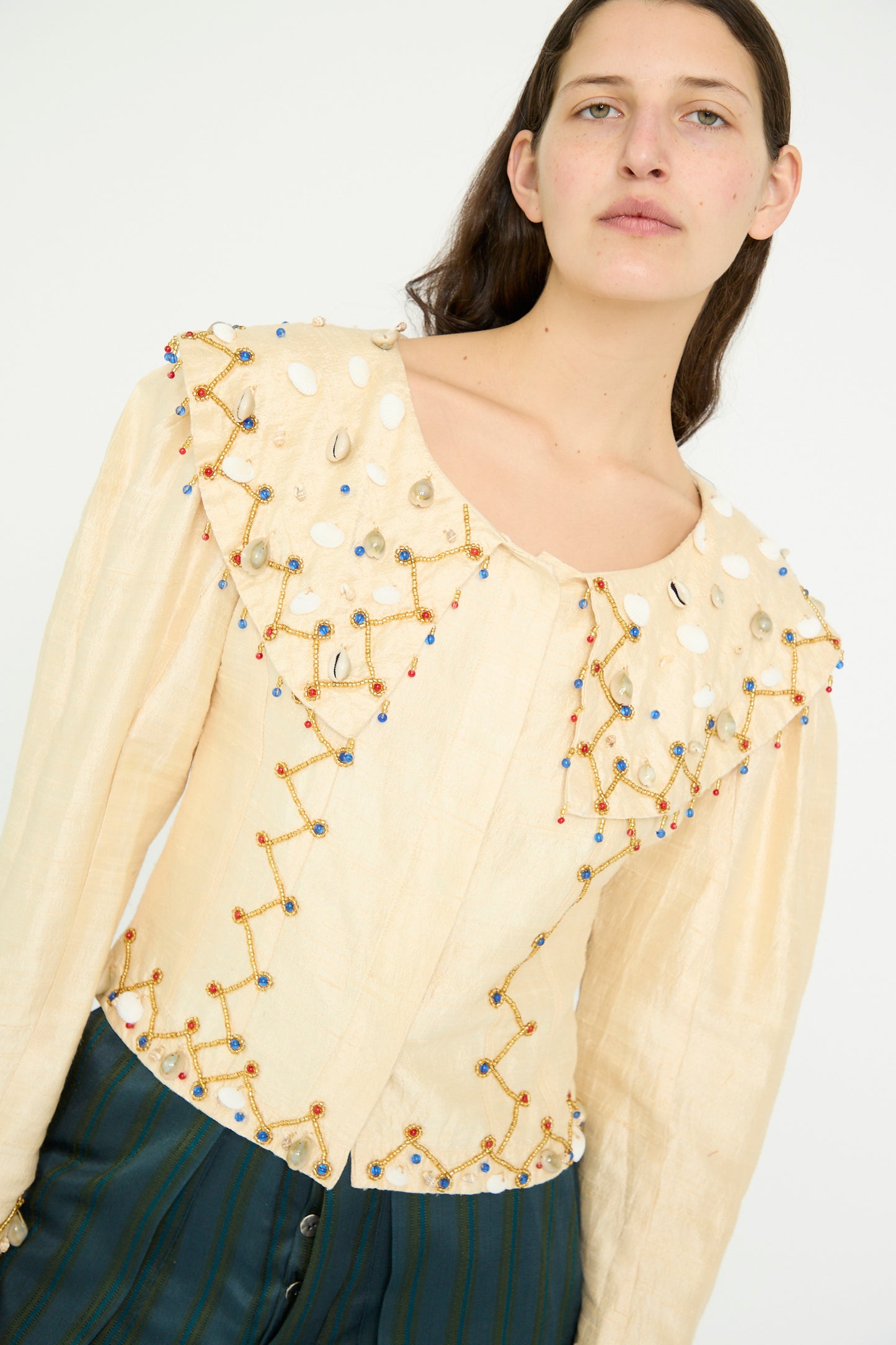 A model stands against a plain background wearing TIGRA TIGRA's Raw Silk Pirate Top in Cream with Gold, featuring embroidered bead designs and cowrie shells, paired with dark green striped pants.