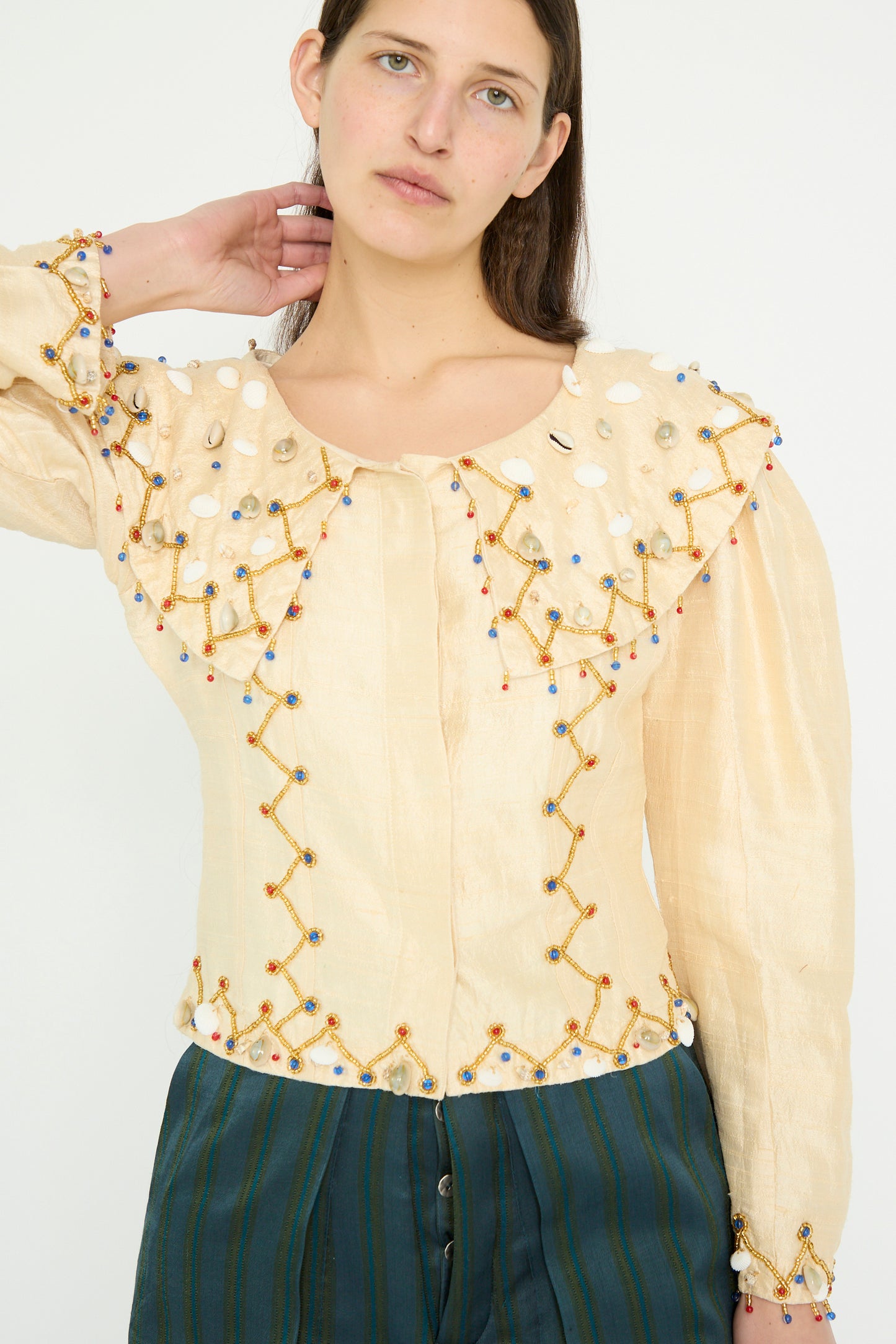 A model poses with one arm raised against a plain backdrop, wearing the TIGRA TIGRA Raw Silk Pirate Top in cream with gold cowrie shells and green-striped pants.