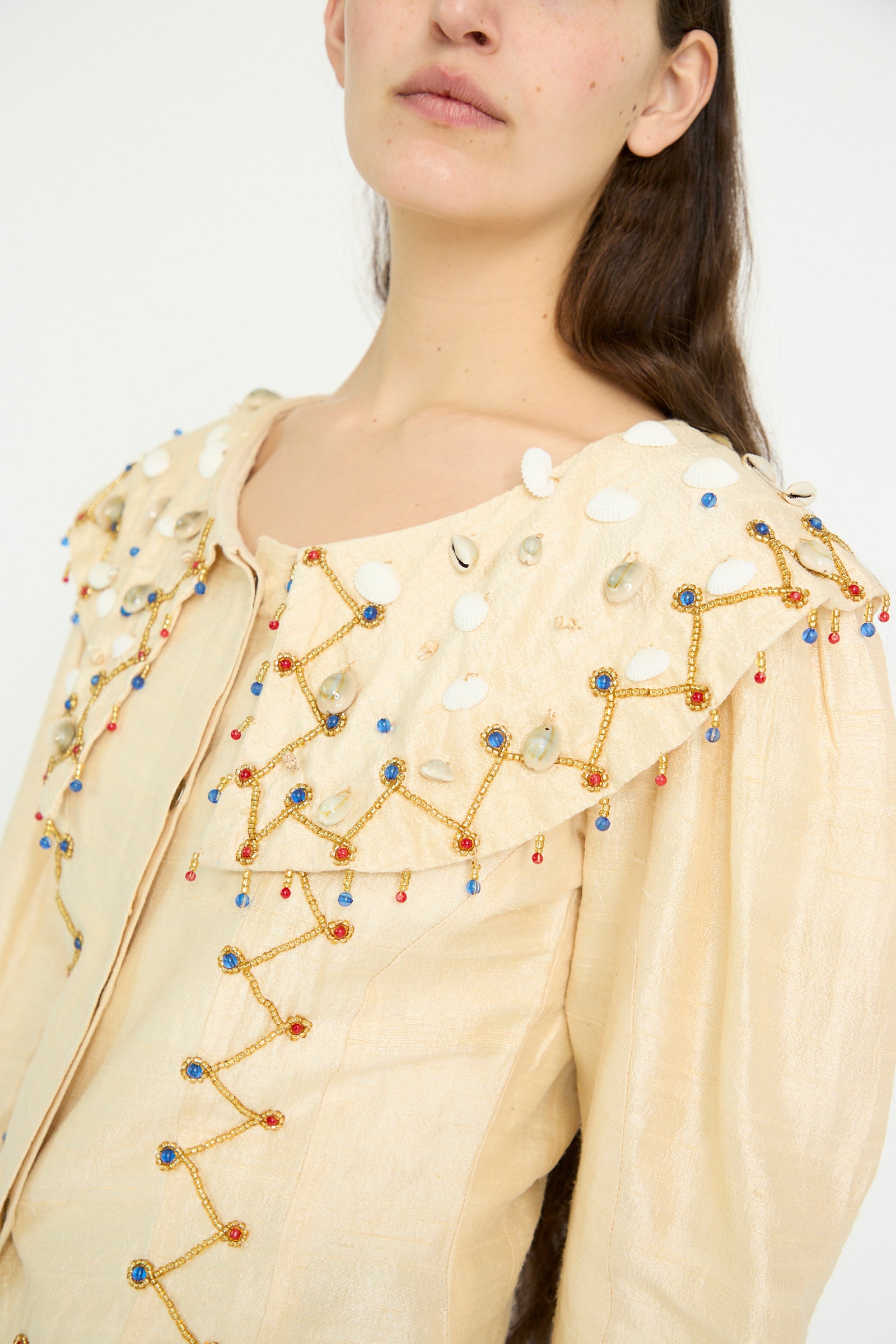 A model wearing TIGRA TIGRA's Raw Silk Pirate Top in cream, featuring cowrie shells, beads, and intricate gold stitching against a plain backdrop.