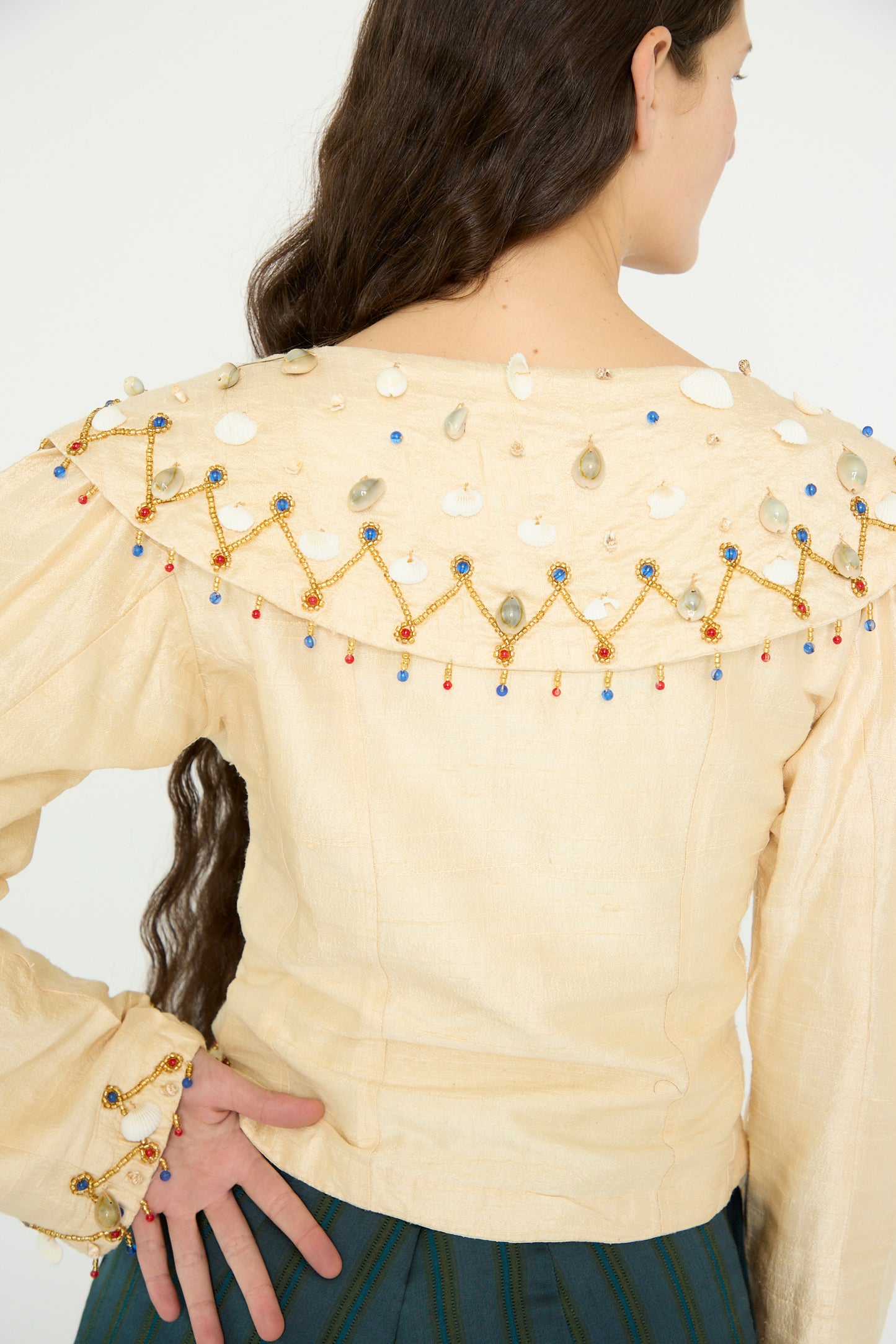 A model wears the TIGRA TIGRA Raw Silk Pirate Top in Cream with Gold, featuring intricate beadwork and cowrie shells on the shoulders and cuffs. Their long brown hair cascades down as they stand with their back to the camera, paired with a dark green pant.