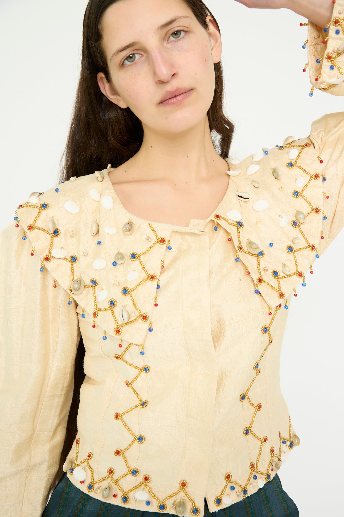 Against a plain background, a person poses with one arm raised, highlighting the Raw Silk Pirate Top in Cream with Gold by TIGRA TIGRA, featuring intricate embroidery, beaded patterns, and cowrie shells.