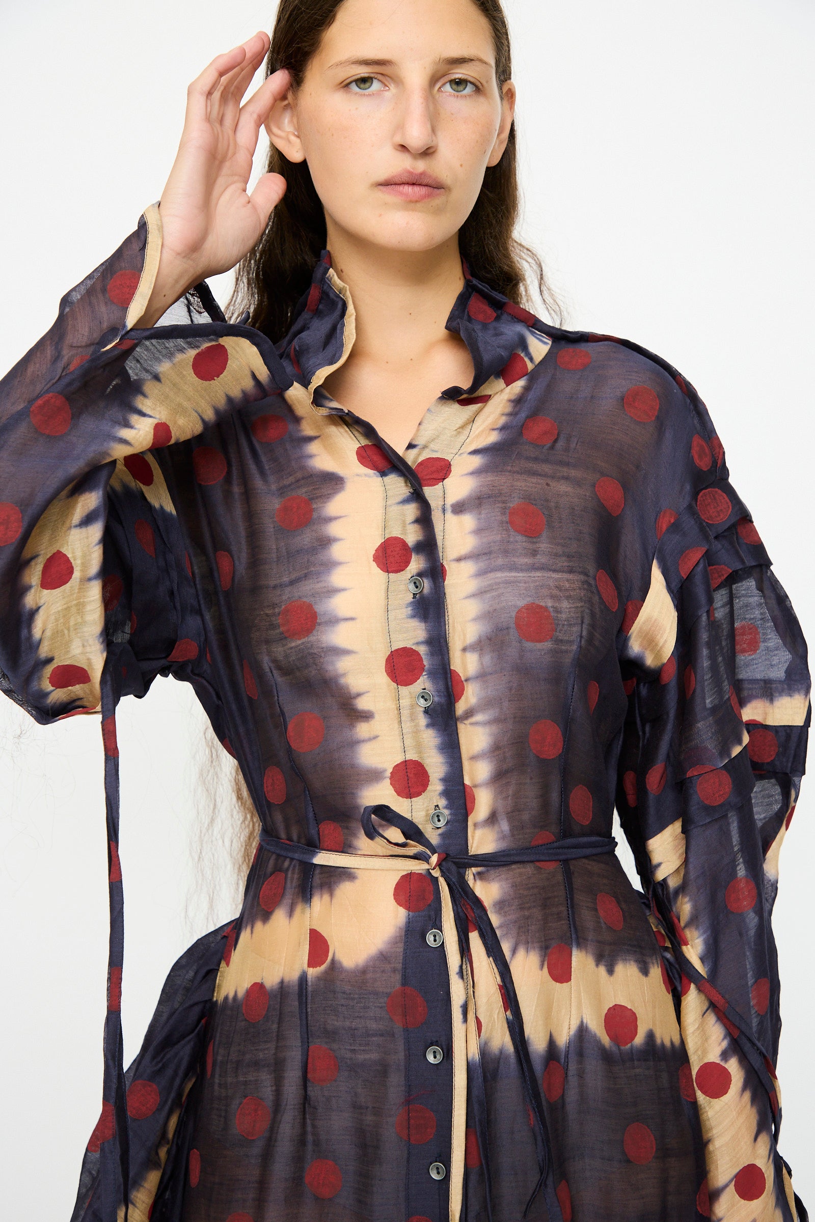 A woman wearing the TIGRA TIGRA Silk Chanderi Teatime Dress in navy, cream, and red—featuring sheer fabric with polka dots, subtle tie-dye patterns, front buttons, and a slim waist tie—stands against a plain background.
