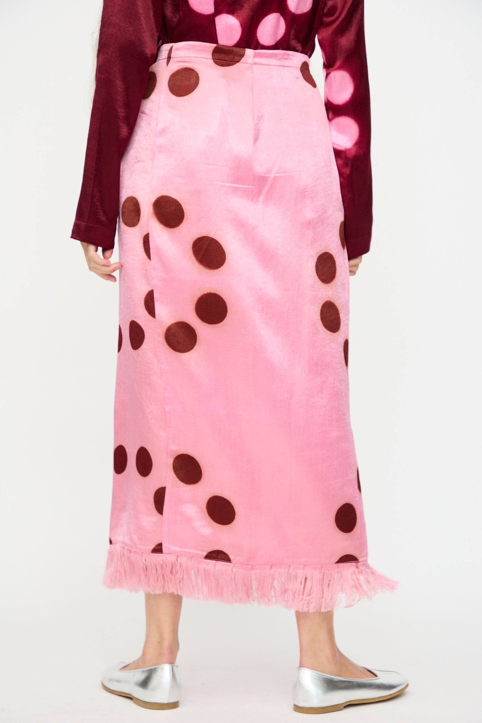 A person shown from the back wears the TIGRA TIGRA Silk Mashroo Moon Dye Wrap Skirt in Pink, featuring large brown spots and fringe at the hem, paired with a maroon top and silver flats.