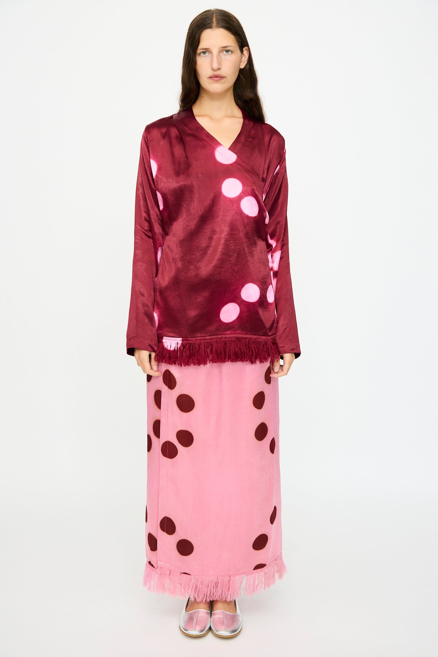 A person stands against a white background wearing the TIGRA TIGRA Silk Mashroo Moon Dye Wrap Top in Maroon, paired with a pink skirt with maroon dots, fringe details, and pink shoes.