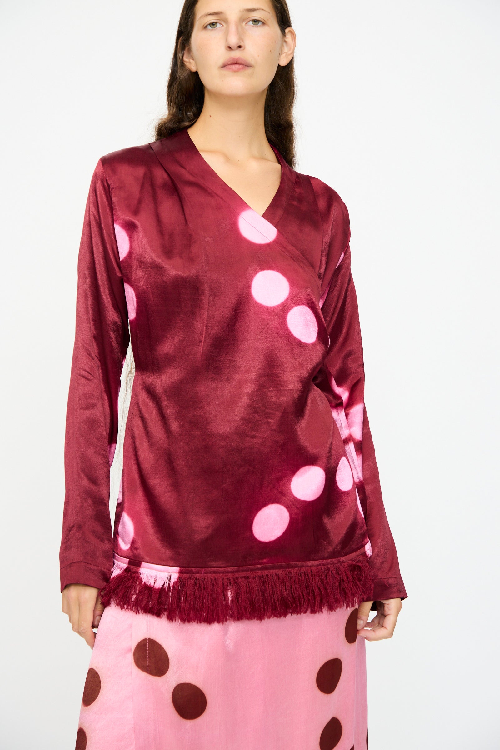 A person wears the TIGRA TIGRA Silk Mashroo Moon Dye Wrap Top in Maroon, featuring fringe trim and a polka dot pattern, paired with a pink skirt with brown polka dots, standing against a plain background.